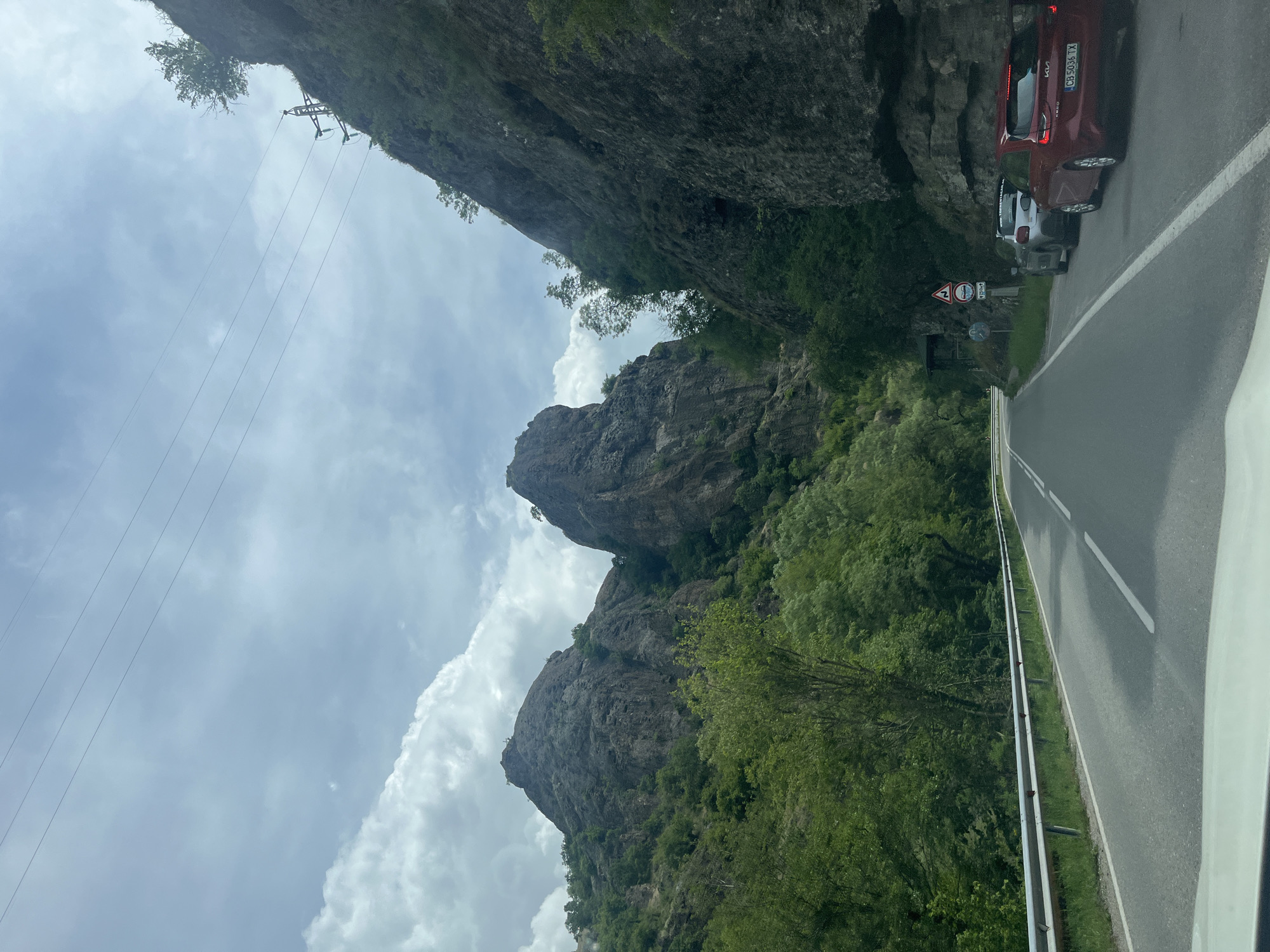 The image depicts a road winding through a mountainous landscape in Rila, Bulgaria. The road is paved and has a metal guardrail. Two cars are parked on the side of the road: a red KIA with license plate CB 9096 TX and a silver car. Road signs are visible near the parked cars, indicating road conditions. The foreground shows the interior of a vehicle from which the photo was taken. The landscape features steep rocky cliffs and lush green trees and vegetation. Overhead, power lines stretch across the sky, which is partially cloudy. The mountain cliffs are rugged and gray, suggesting natural rock formations.