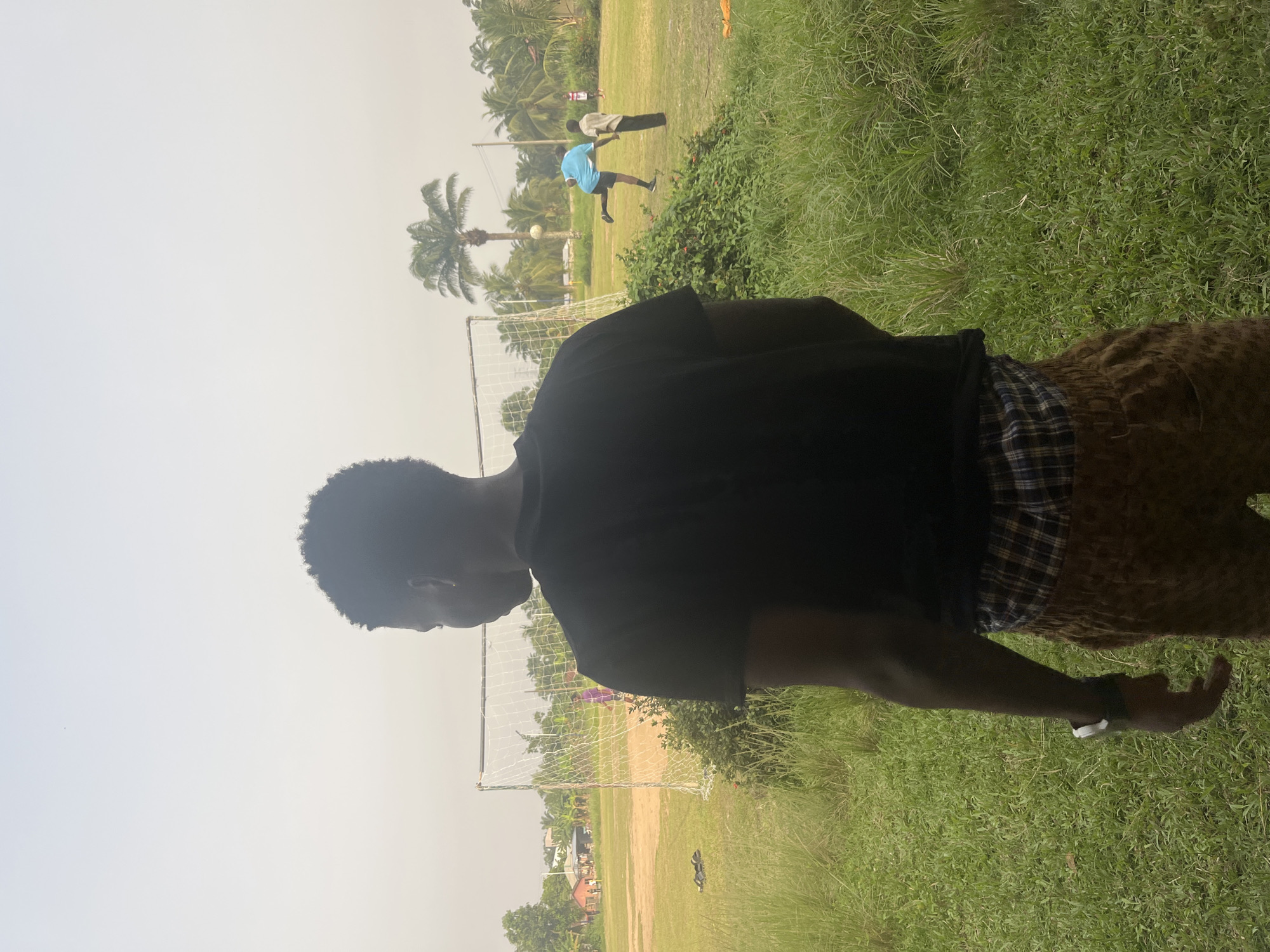 A person is standing on a grassy field near a soccer goalpost in Ikot Ekpene, Nigeria. Another person is seen kicking a soccer ball on the same field. The sky is overcast. Palm trees are present in the background.