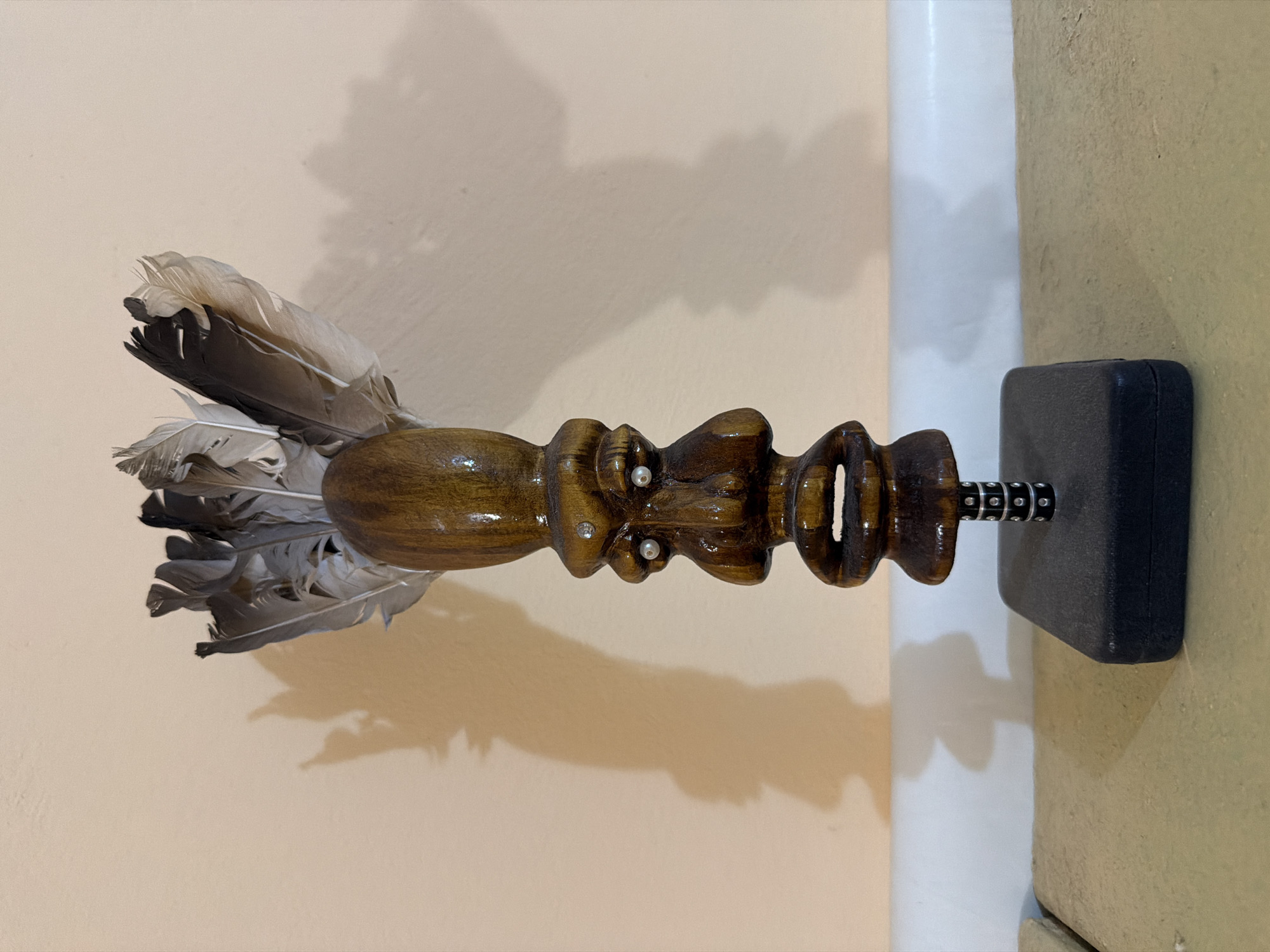 This is a close-up, eye-level, full shot of a carved wooden sculpture on a black base. The sculpture is of a stylized face with two pairs of eyes, an elongated nose, and exaggerated lips. It is adorned with feathers on its head and is mounted on a metal rod with decorative bands. The sculpture is placed in front of a pale peach-colored wall that casts a shadow of the object. The base of the sculpture rests on a textured, sandy-colored surface.