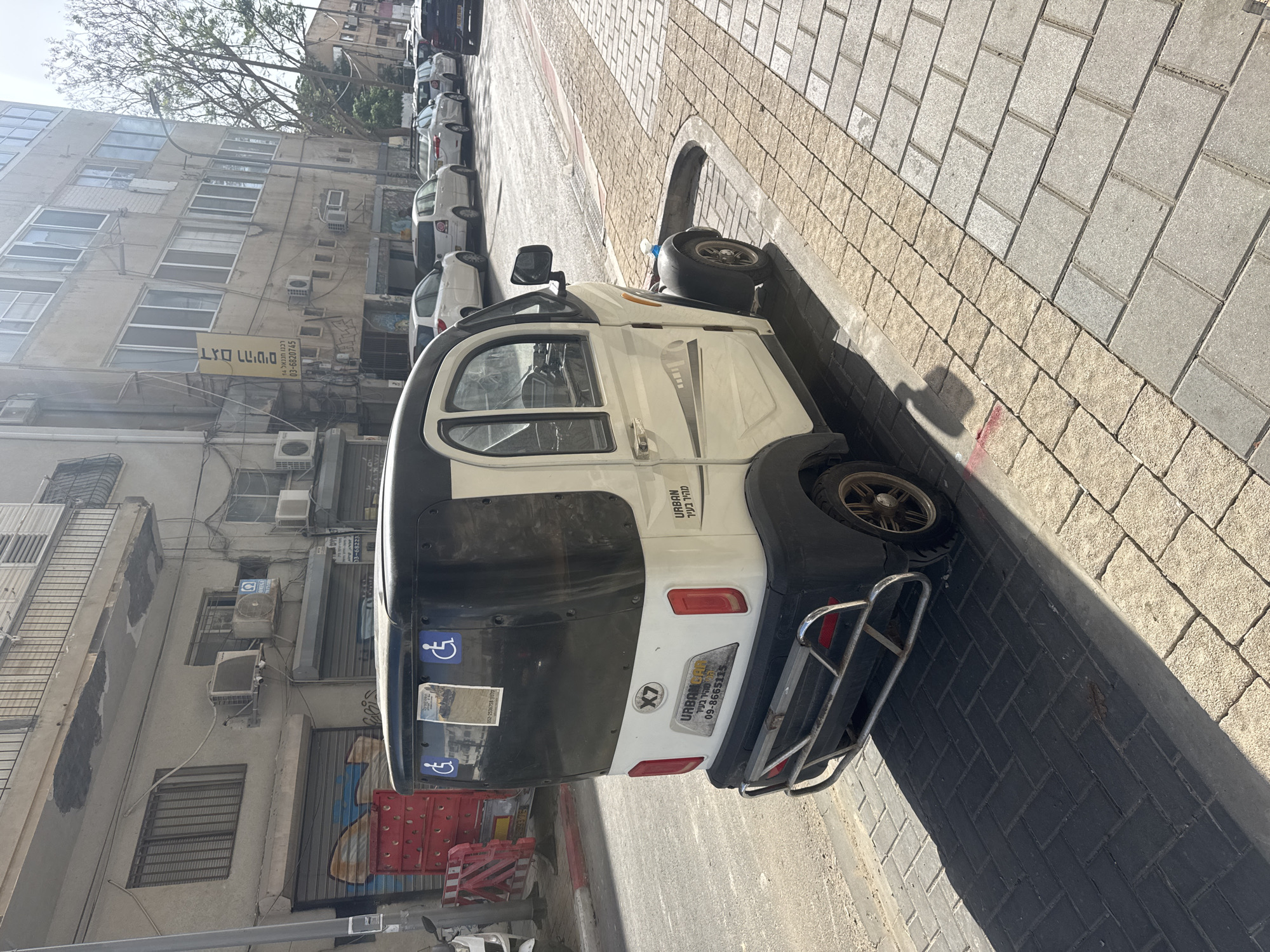 The image captures an Urban Car, a compact three-wheeled vehicle, parked on a brick-paved street in Tel Aviv, Israel. The vehicle is white with black accents and features accessibility stickers. The surrounding environment consists of residential buildings with visible air conditioning units, parked cars along the street, and commercial signs in Hebrew. The scene appears to be a typical urban street setting.