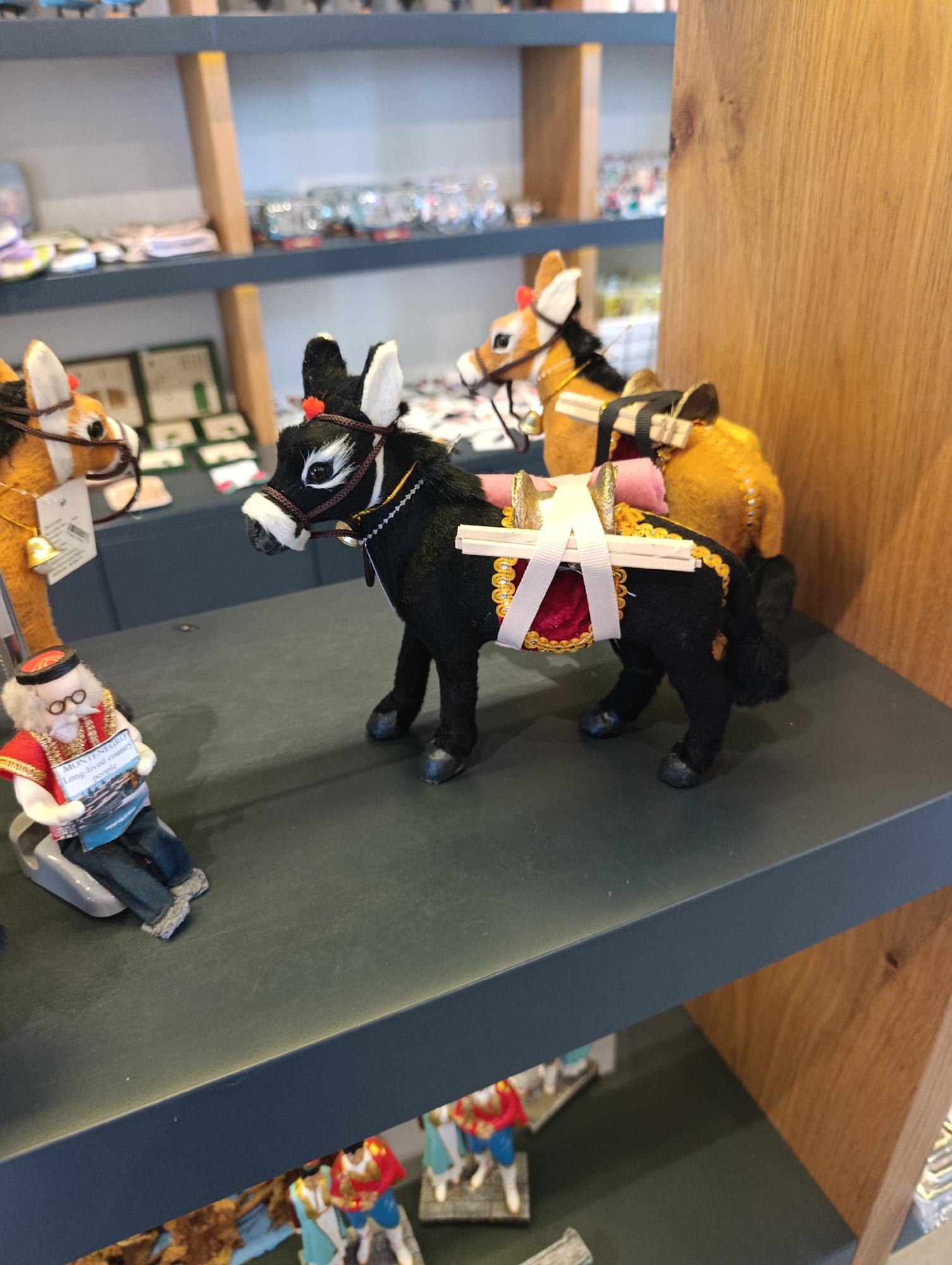 In a shop display in Kuk, Montenegro, two stuffed donkey figurines are positioned on a dark gray shelf. The donkey in the foreground is black and has a saddle with red and gold trim, adorned with white ribbon and wooden dowels. A small, seated figurine of an elderly man wearing a red jacket, glasses, and a traditional cap is placed to the left of the donkey, holding a brochure. Behind the black donkey, a second, tan-colored donkey figurine is visible, also with a saddle. The background shows shelves with various items, suggesting a souvenir or gift shop.