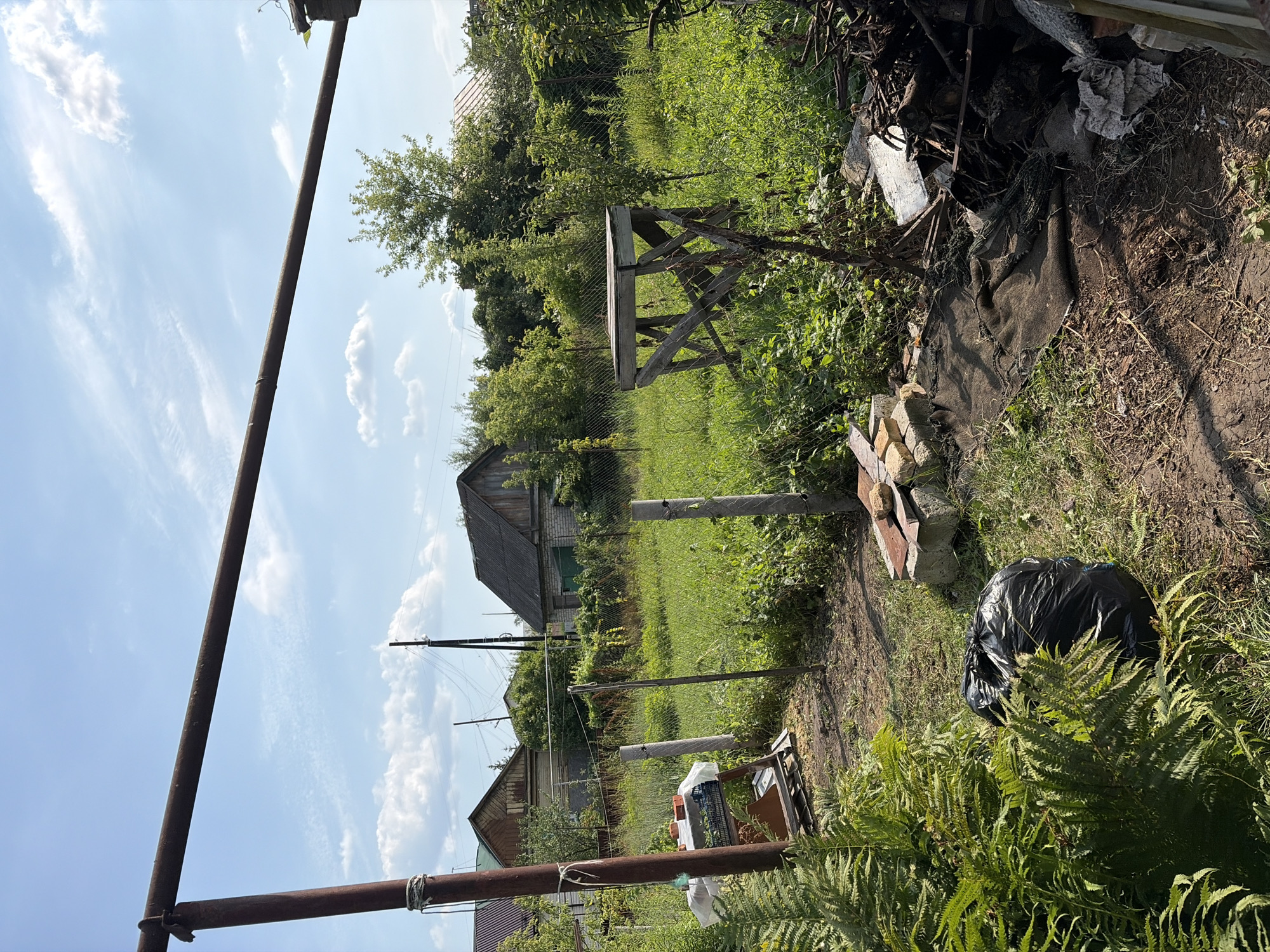 This is a ground-level, eye-level, medium shot of a rural backyard in Tambov, Russia, on a sunny day with scattered clouds. A rusty metal bar stretches horizontally across the top of the frame, supported by a red-brown vertical pole on the left. The backyard is overgrown with tall green grass and various plants, including ferns in the foreground left.
Several structures are visible. On the left, a makeshift table with a white tarp is set up next to the vertical pole, with a plastic basket and some bricks on it. Two concrete posts stand in the yard, one near the table and another further back. A wooden structure with an X-frame is situated in the middle ground right.
In the background, there are several houses with visible roofs and walls. A chain-link fence runs through the yard, separating different areas. A pile of debris, including wood and branches, is located in the bottom right corner. A large black trash bag is partially obscured by ferns in the bottom left. Power lines and poles are visible in the distance. The overall impression is one of a somewhat unkempt, lived-in outdoor space.