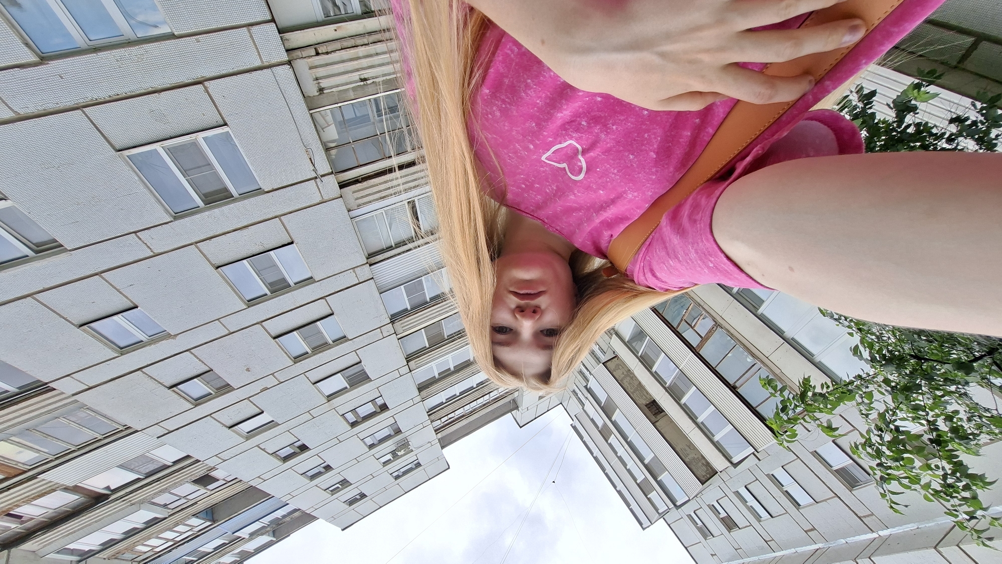 A young woman with long blonde hair is taking a selfie in front of a residential building in Chaikovsky, Russia. She is wearing a bright pink t-shirt with a white heart graphic on it and a brown strap from a bag crosses her chest. The building behind her is a typical Soviet-era apartment block, with rows of windows and balconies. The sky is overcast. The perspective is a low-angle shot, making the building appear larger and more imposing.