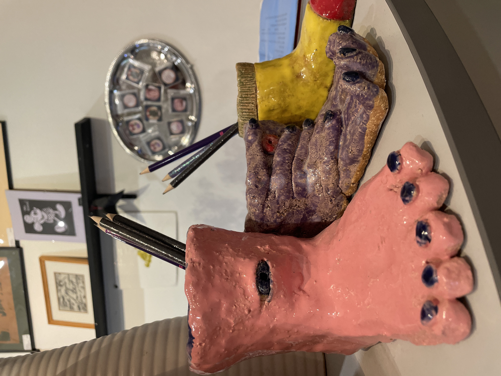 This is an image of an art installation, featuring three stylized, ceramic feet in pink, purple, and yellow hues. Pencils are inserted into holes in the pink and purple feet. The feet are positioned on a grey surface. In the background, there's a display of framed artwork and a silver tray containing small, square images. The image does not provide any information related to the city of Khimki, Russia.