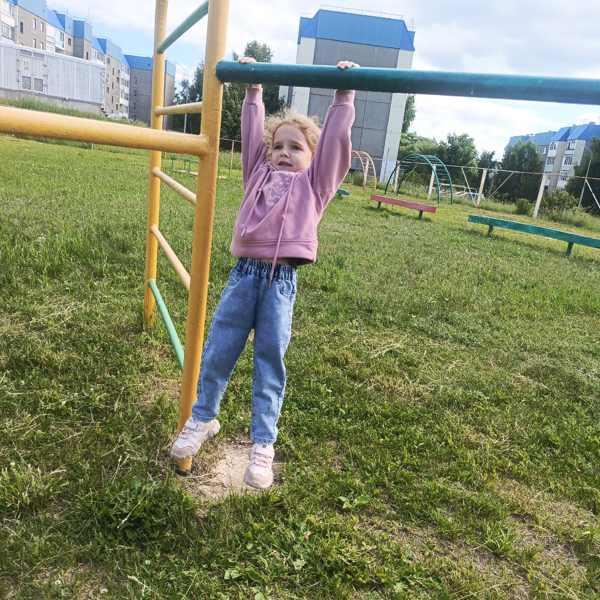A young Caucasian girl with curly blonde hair, wearing a pink hooded sweatshirt and blue jeans, is hanging from a green horizontal bar on a playground structure. The playground has yellow supports. The girl is looking towards the camera with a neutral expression. The background shows a grassy field, trees, and multi-story apartment buildings with blue roofs. The scene appears to be located in a residential area, possibly in the city of Markovskii, Russia, given the architectural style of the buildings and the vegetation. The weather is overcast with some patches of blue sky.