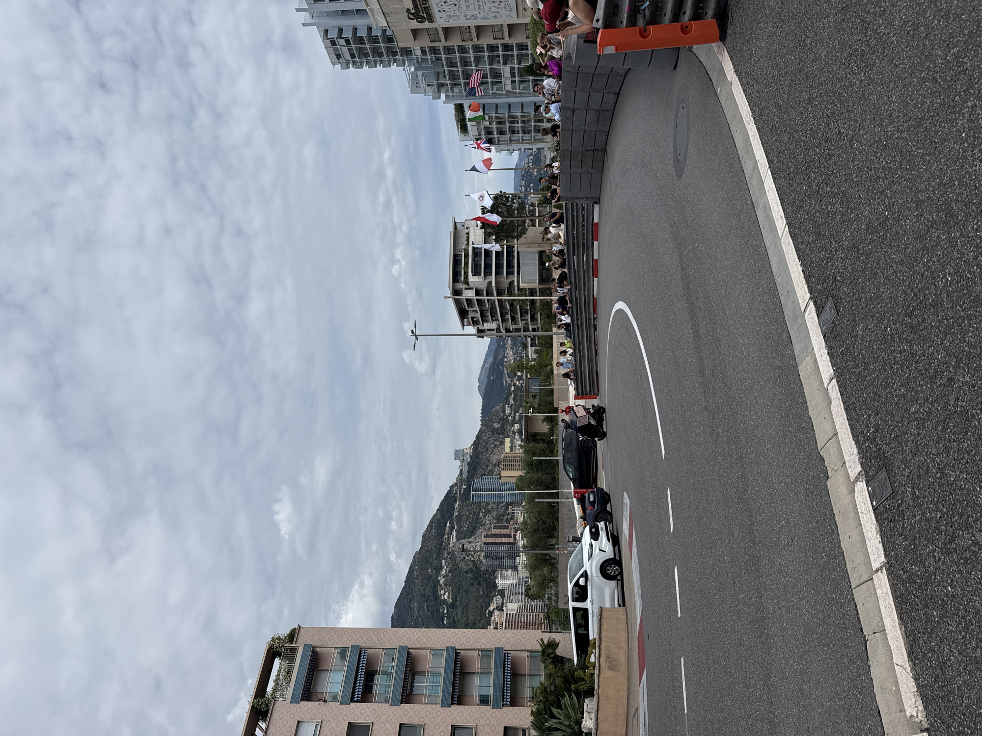 The image shows a street scene in Monaco. A curved road is visible, lined with concrete barriers on one side and buildings on the other. People are standing on the buildings watching the road. Several cars are parked along the roadside. Beyond the buildings, a hillside covered with more buildings can be seen, leading up to a mountain ridge. The sky is overcast.