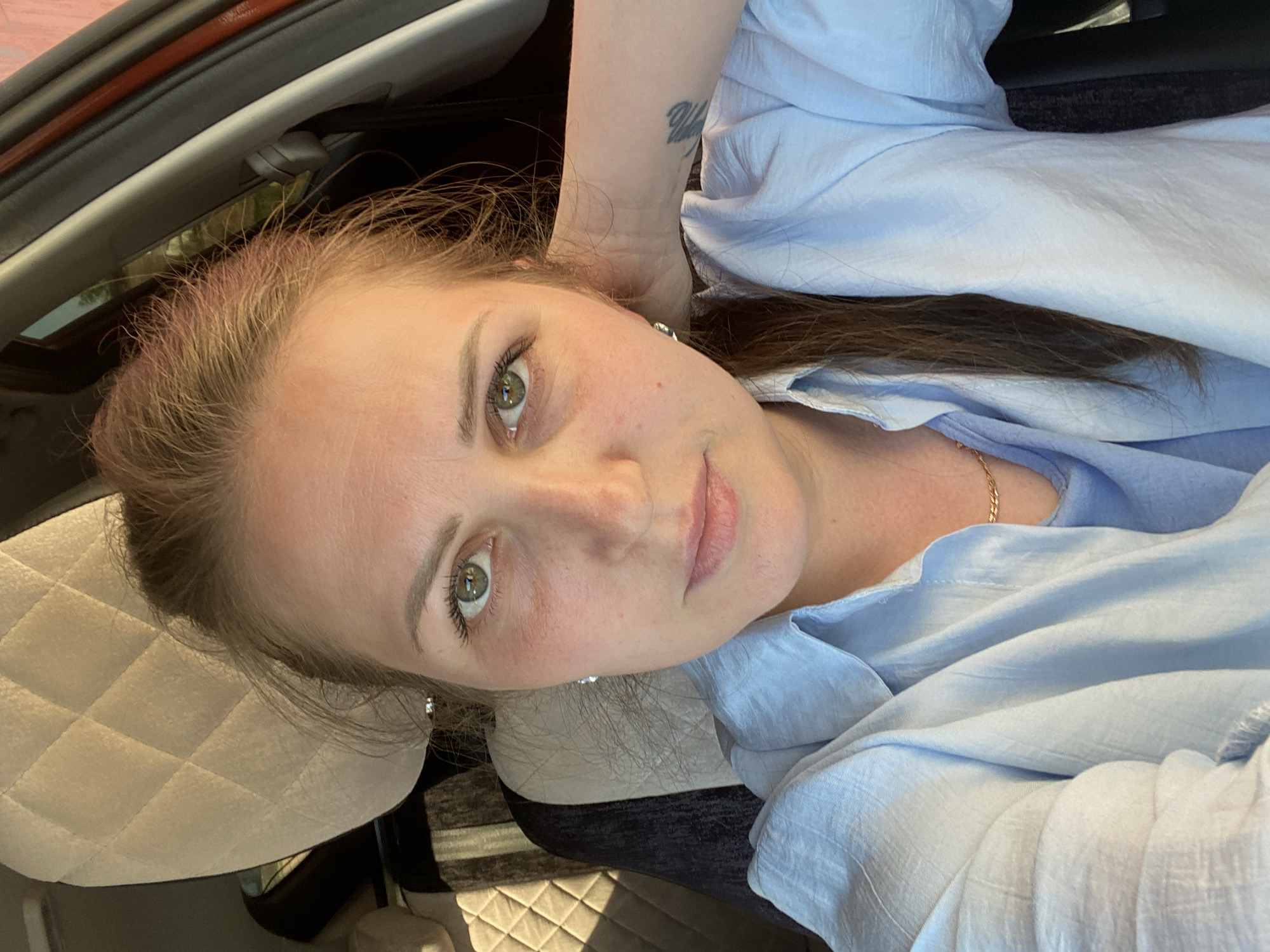 A woman with fair skin, green eyes, and brown hair is shown in a close-up shot. She is wearing a light blue blouse. Her head is resting on a light-colored, quilted surface that appears to be a car seat. Her left arm is raised, with a tattoo visible on her wrist. The background includes part of the car interior, including the roof and window frame. The location in the city of Markovo, Russia, cannot be determined from the image.