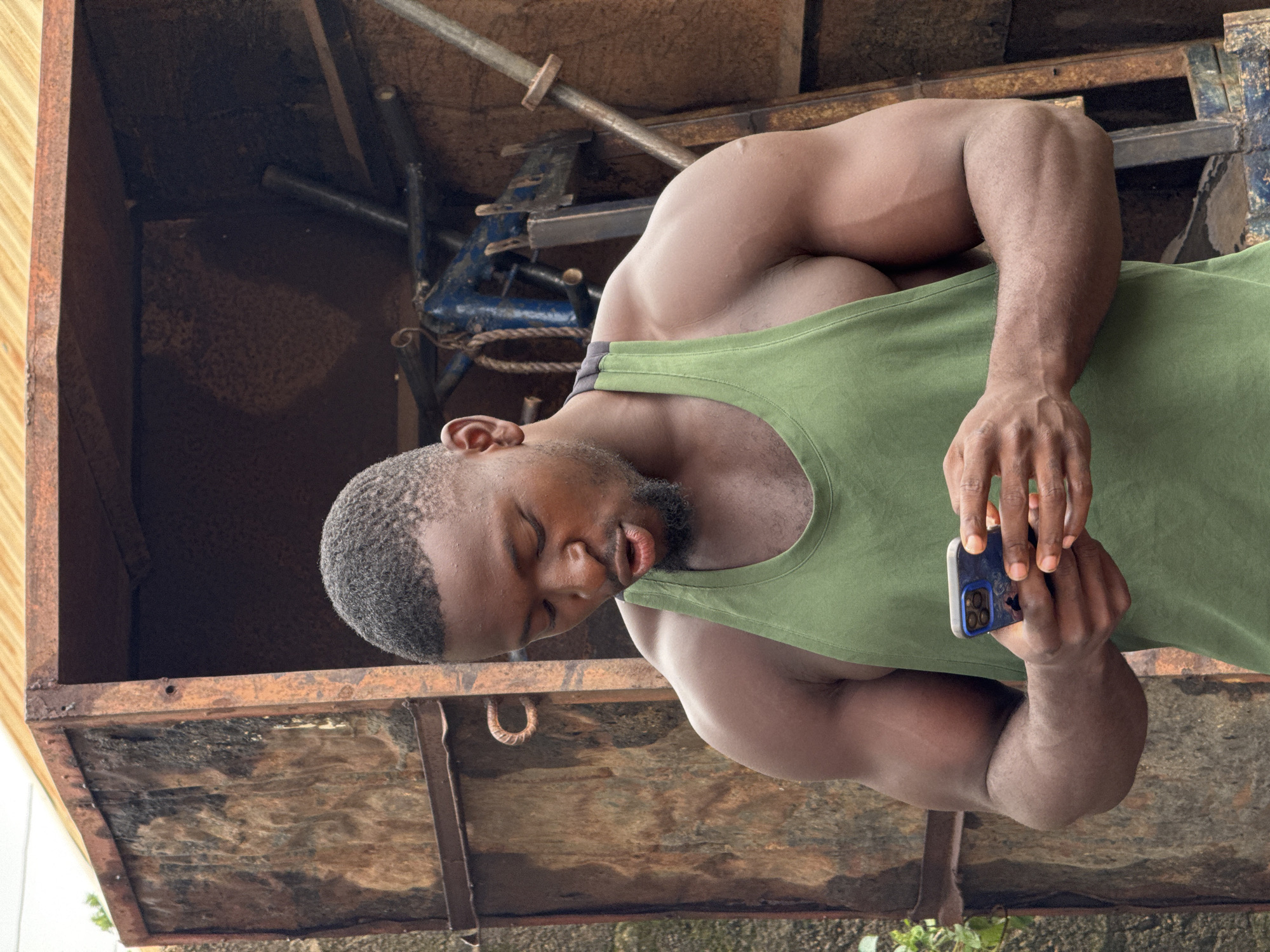 In this media file, a muscular Black man is pictured from the chest up, holding a smartphone in his right hand. He is wearing a green tank top, and his head is tilted down, with his eyes closed. His skin appears to be lightly dusted with what could be chalk or sweat, suggesting a recent workout.
The background is industrial and somewhat weathered, with a wooden and rusty metal structure visible. The lighting suggests it could be daytime, possibly overcast, as there are no harsh shadows. The overall scene appears to be in a gym or a workshop environment. The man's expression is relaxed, perhaps weary or focused.
There is no visible text in the image. The context provided places this scene in Akwanga, Nigeria.