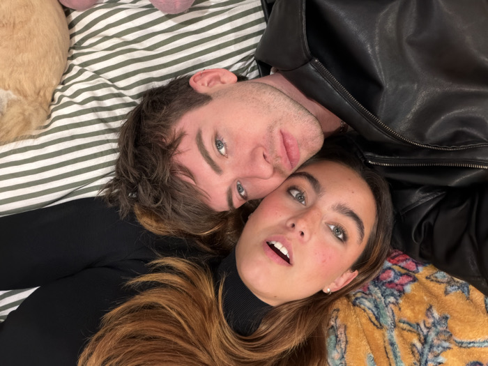 A young man and woman are lying side-by-side on a bed. The man has short brown hair and blue eyes, while the woman has long brown hair and light eyes. She wears a black turtleneck and small earrings. The man is wearing a black leather jacket. They are lying on top of a bed with striped sheets and a yellow floral-patterned blanket. The couple appears relaxed and casual.