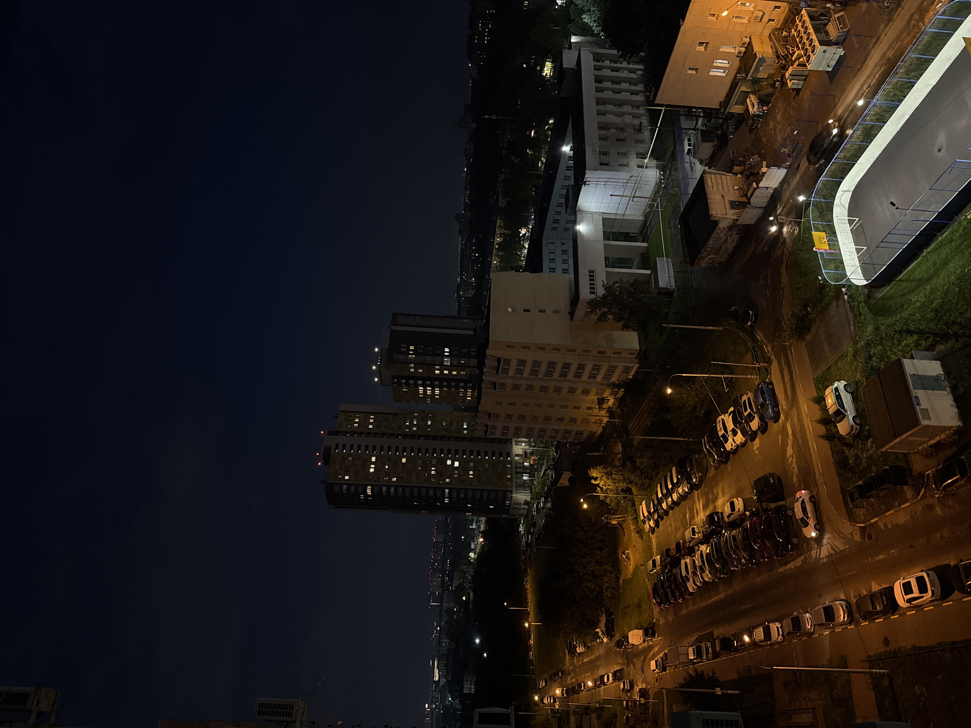 This is a night-time aerial photograph taken in Nizhny Novgorod, Russia, likely from a high-rise building. The scene depicts a residential area with a street lined with parked cars, a dark forested area, and several apartment buildings of varying heights. In the foreground, a sports court, possibly a mini-football or basketball court, is visible with blue fencing and a yellow hoop. The sky is dark and overcast, with only a few distant lights indicating the presence of other urban areas or industrial sites. The lighting in the image is predominantly from streetlights and the illuminated windows of the buildings, casting a warm glow on the surroundings. There are no people visible in the photograph.