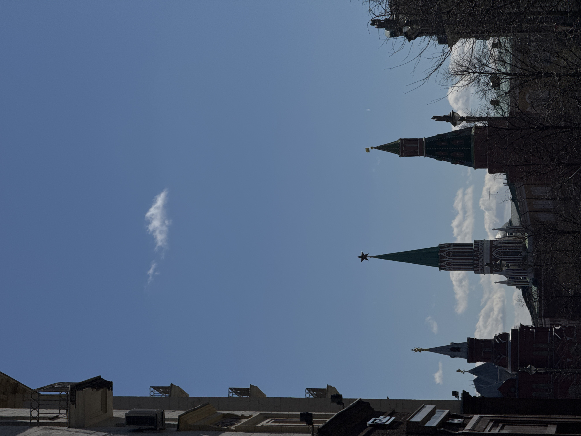 The image presents a view looking up towards a clear blue sky, with a single, elongated white cloud. The lower and upper portions of the frame contain architectural elements and bare tree branches.
In the lower part, building rooftops with miscellaneous rooftop fixtures like vents or air conditioning units are visible. The upper part of the frame reveals the tops of several towers of the Moscow Kremlin. These towers have distinctive architectural designs with pointed roofs, decorative elements, and one is topped with a star. Bare tree branches obscure some of the towers. The environment appears to be a sunny day, and the picture conveys a sense of looking up from street level in Moscow.