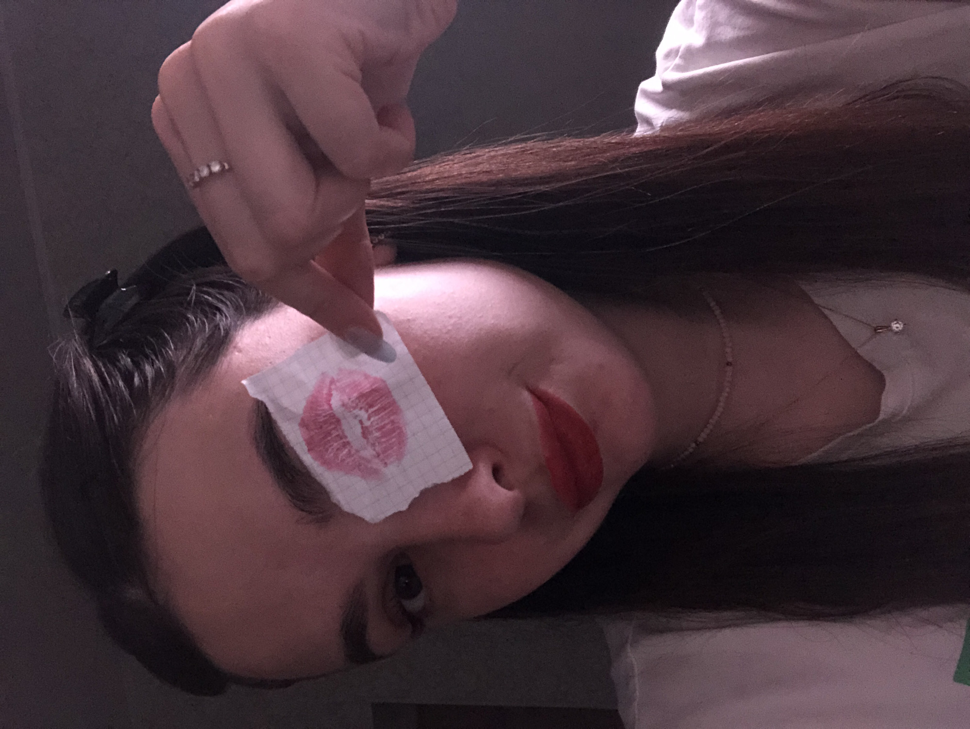 The image shows a woman with dark hair and red lipstick, lying on her side. She is holding a small piece of paper with a lipstick kiss print on it near her eye. She is wearing a ring on her finger and a beaded necklace with a pendant. The background is blurry and indistinct, but appears to be an indoor space. The location in the city of Noviy, Russia is not discernible from the image.