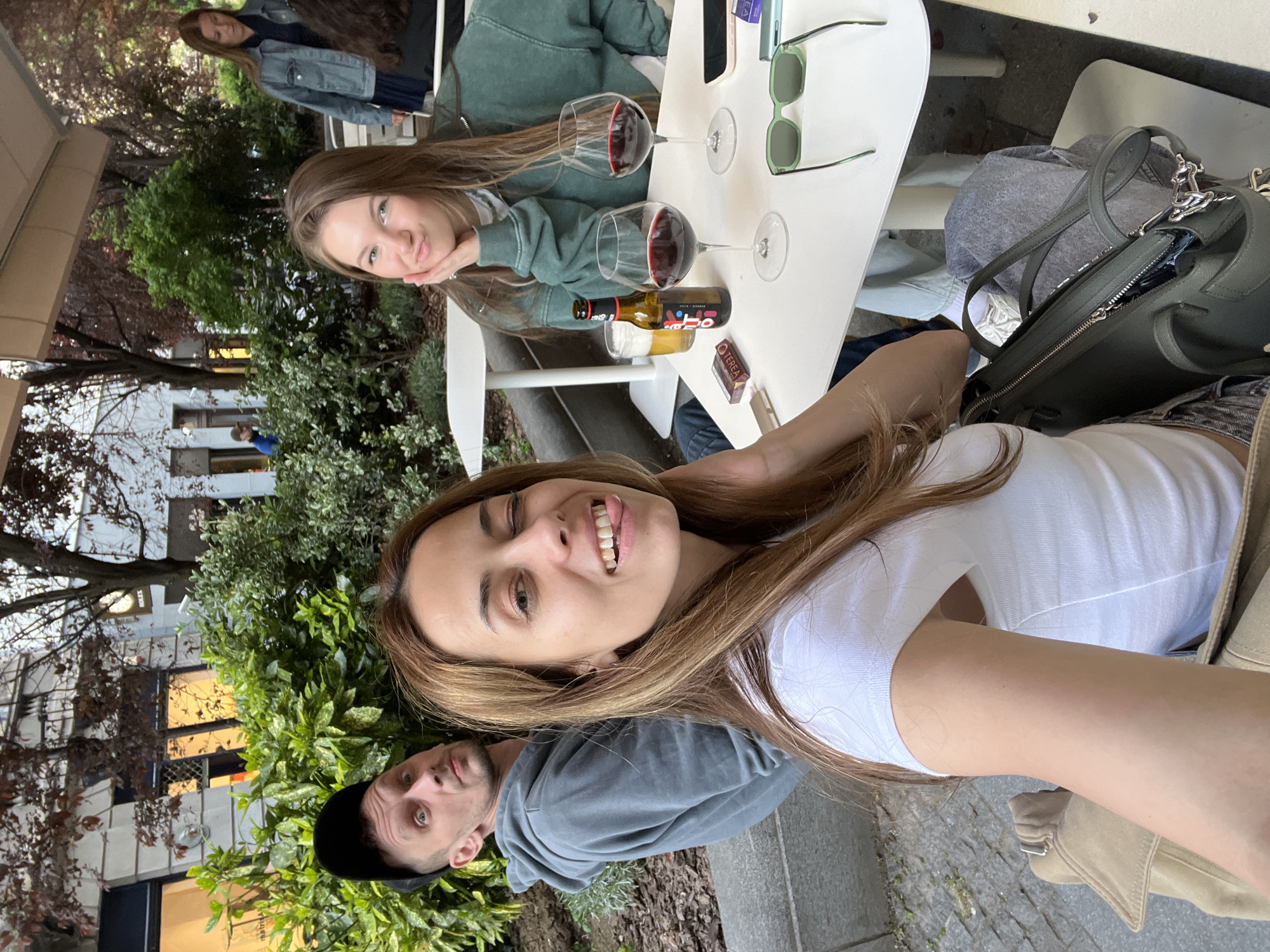 An outdoor, rotated photograph captures a social gathering of four individuals, likely friends or family, enjoying drinks at what appears to be a cafe or restaurant patio in Belgrade, Serbia. The image is oriented with the top of the frame to the right.
In the immediate foreground, a young woman with long brown hair is smiling broadly at the camera, her tongue slightly visible. She wears a white t-shirt, and a beige jacket is draped beside her. To her left, a man in a dark grey t-shirt and black cap looks at the camera with a neutral, slightly surprised expression, partially obscured by greenery.
At a white table in the mid-ground, another young woman with long brown hair, wearing a green hoodie, leans forward with a subtle smile. On the table are two wine glasses filled with red wine, a bottle (possibly beer or wine, with partially legible text), a smartphone, a pair of green-framed sunglasses, and a pack of "TEREA" brand cigarettes. Next to the foreground woman is a dark green handbag.
Further in the background, a fourth person, a woman in a denim jacket over a dark top, is partially visible, also looking towards the camera. The setting is characterized by lush green bushes and trees, with urban buildings in the background. The lighting suggests it is daytime, possibly late afternoon or early evening, and the mood is relaxed and convivial.