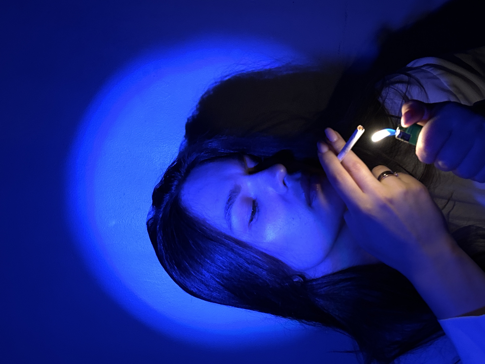 In a dimly lit indoor setting, likely a residential room in Chaikovsky, Russia, a young woman with long dark hair is captured in the act of lighting a cigarette. She is illuminated by a vibrant blue light, creating a stark contrast with the shadows. Her eyes are cast downward, focused on the cigarette and the green lighter in her hand, which emits a bright flame. A circular halo of the blue light is visible on the textured wall behind her. She wears a white garment, and a ring is present on her left ring finger. The overall atmosphere is moody and intimate, with the blue lighting casting an ethereal glow.