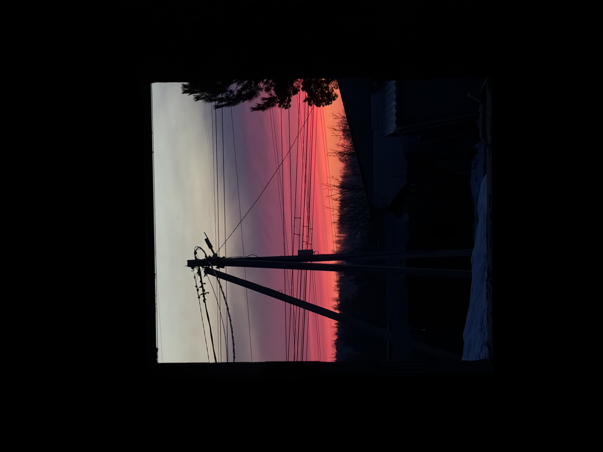 The image, shot from inside a room through a window, features an electrical pole and wires silhouetted against a vibrant sunset sky. The sky displays a gradient of colors, transitioning from light gray near the horizon to pink and red hues higher up. The silhouettes of trees and possibly rooftops are visible on the right side of the frame. The scene appears to be set in a residential area, with power lines suggesting a suburban or rural context. Snow is visible to the bottom right of the window. While there is no conclusive evidence, the information given indicates it may be the city of Chaikovsky, Russia. There are no people visible in the image.