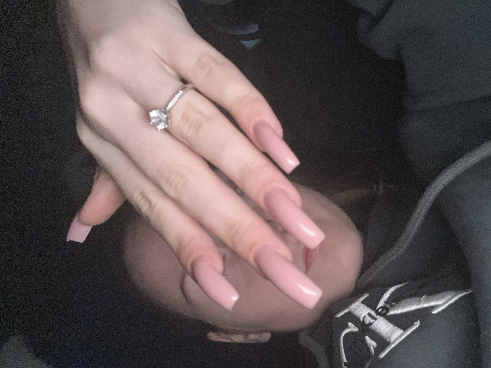 A close-up shot captures a person's left hand with long, light pink, square-shaped artificial nails. The hand has a light skin tone and wears a silver ring with a prominent, round, clear stone on the ring finger. The hand is positioned over the lower part of a person's face, partially obscuring the nose and mouth area, which are slightly out of focus.
The background is dark and indistinct, suggesting an indoor or dimly lit environment. The person's face beneath the hand appears to be resting. The individual is wearing dark clothing, likely a hoodie or sweatshirt, visible on the right side of the frame. On this dark fabric, there is a white embroidered, stylized logo or letter, characterized by angular elements. While the provided context indicates the location is Varna, Bulgaria, there are no specific visual cues within the image to confirm this. The overall mood is calm and quiet, with no explicit emotions visible.