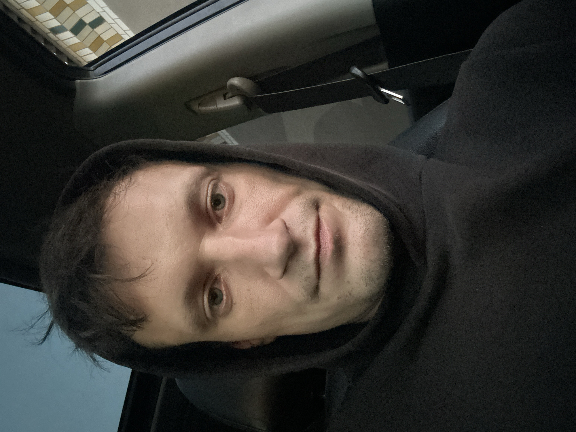 A close-up, medium-shot selfie shows a fair-skinned man in his late 20s to early 30s looking directly at the camera. He has light brown, messy hair that is slightly tousled and falls over his forehead. He has grey-green eyes, a straight nose, and thin lips. His face is slightly stubbled. He is wearing a black hooded sweatshirt, with the hood pulled up around his head, obscuring part of his hair and forehead. The man appears to be seated in the driver's seat of a car, as indicated by the presence of a seatbelt mechanism visible in the background to his right. The car's interior is dark, with the exception of a sliver of light coming from the sunroof above him, revealing a pale blue sky. To the right of the man, a portion of the car's interior trim is visible, featuring a decorative pattern of cream, yellow, and teal squares. The scene is indoors, within a vehicle, and does not provide any information about the city of Shatava, Ukraine.