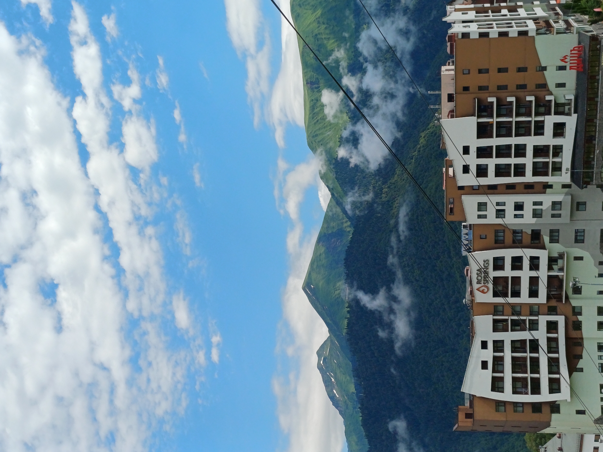 This is a medium shot, eye-level, outdoor photograph capturing a modern resort building in Estosadok, Russia, nestled against a backdrop of lush green mountains partially shrouded in clouds. The sky above is a vibrant blue with scattered white clouds.
The resort building is the primary subject, featuring a distinctive architectural style with white and brown facade panels and numerous windows. A prominent sign on the building reads "ROSA SPRINGS" in orange lettering, with a stylized flame logo. Cables from a ski lift are visible in the foreground, stretching across the frame.
The mountains behind the resort are covered in dense, dark green forest at their base, transitioning to lighter green slopes at higher elevations. Some patches of snow are visible on the mountain peaks. Wisps of white clouds drift amongst the mountain ranges, adding depth and atmosphere to the scene. The overall impression is one of a scenic mountain resort town on a clear, but possibly cool, day.