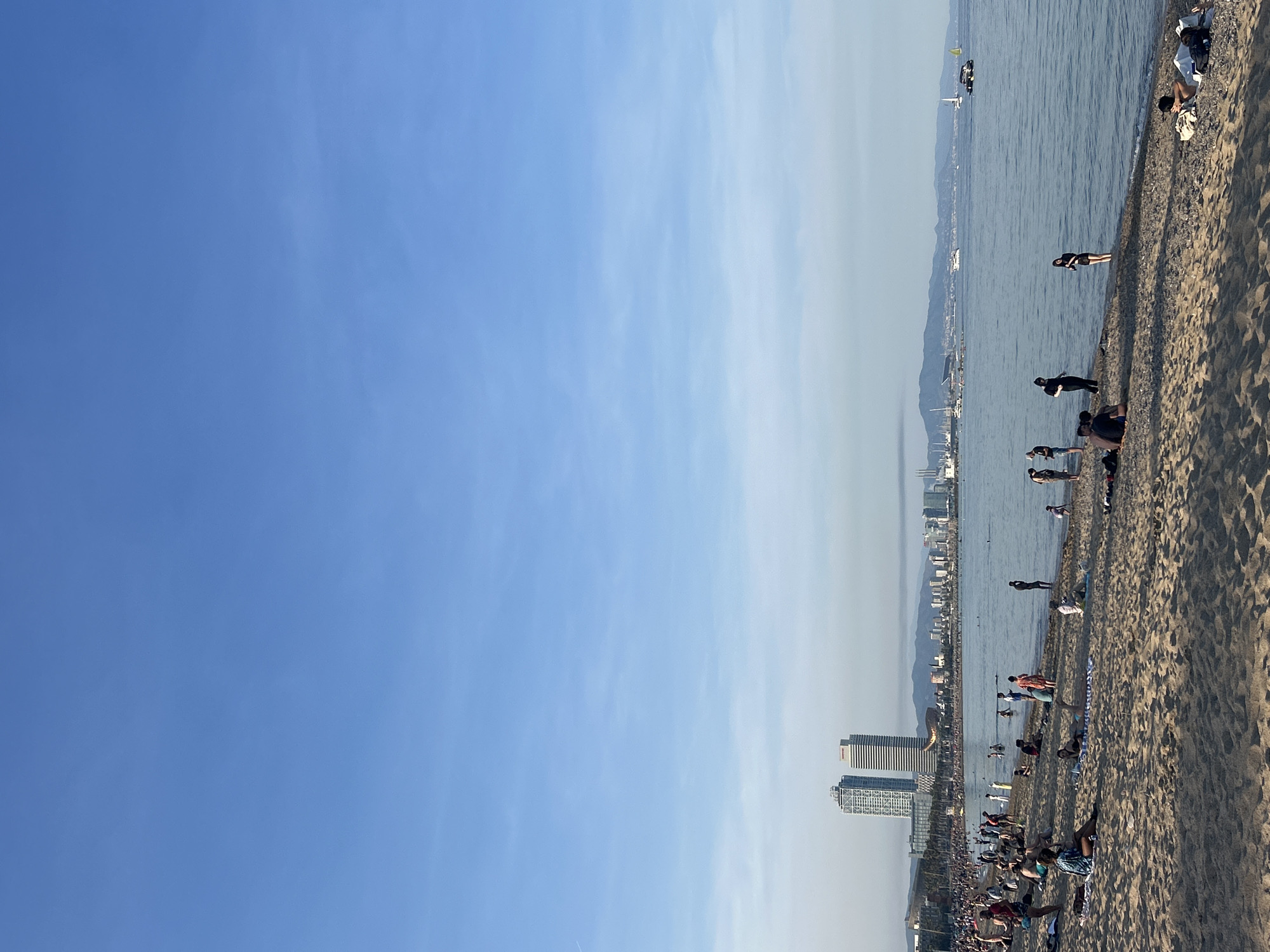 The image depicts a beach scene in Barcelona, Spain. The foreground is dominated by a crowded sandy beach with people sunbathing, walking, and playing. The sea stretches out to the horizon, dotted with boats and swimmers. In the background, the Barcelona skyline is visible, including the iconic Hotel Arts and Torre Mapfre. The sky is a clear blue with light clouds.
