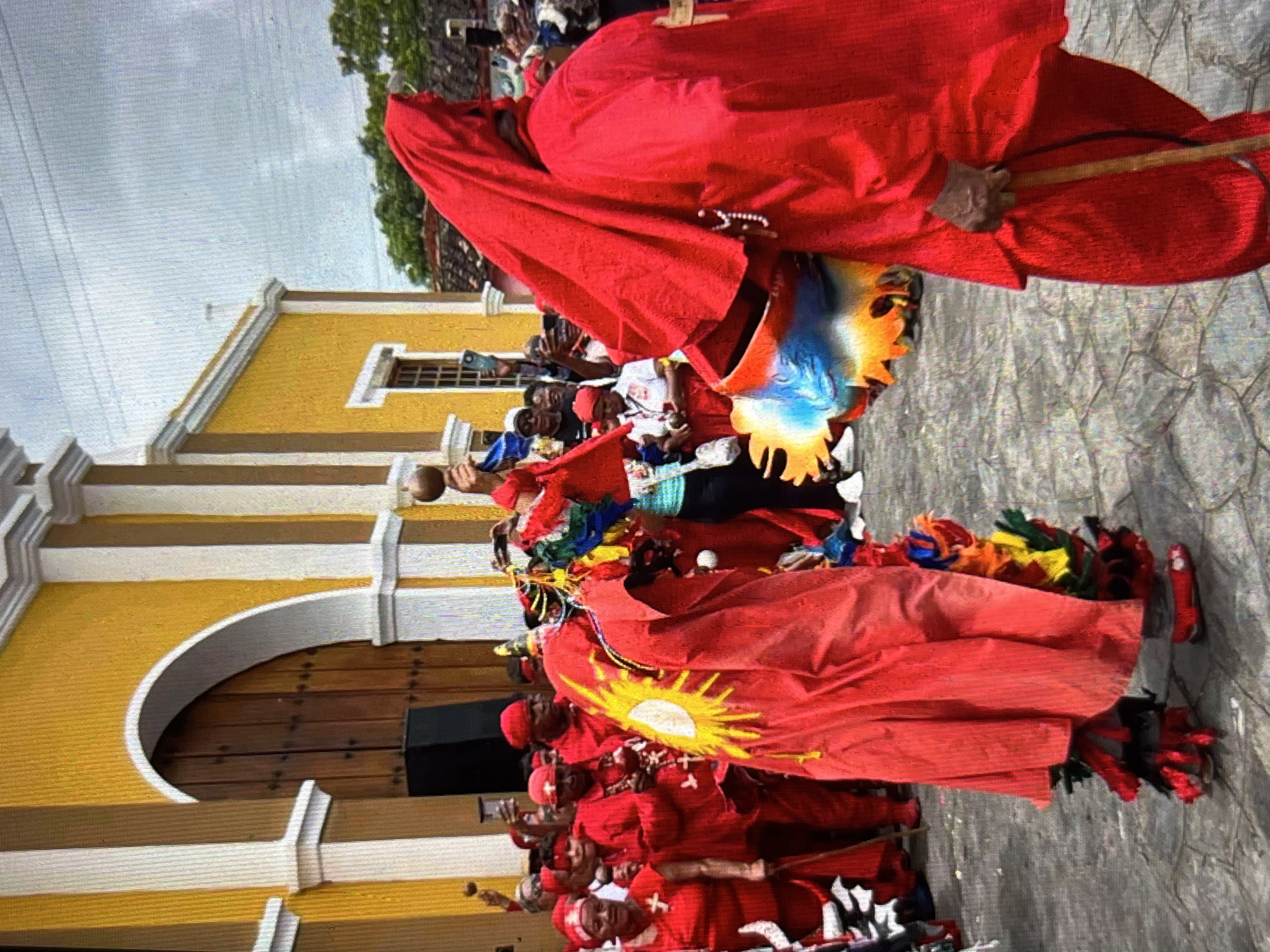 In Tinaquillo, Venezuela, a festive scene unfolds in front of a vibrant yellow church with white and brown columns. The foreground is occupied by two individuals in elaborate red costumes. One figure, facing left, wears a long, flowing red robe adorned with a large yellow sun design and colorful feathered decorations around the ankles. They hold a maraca in their right hand. Facing away from the viewer, the second figure is also clad in a red hooded robe and carries a wooden cane. The costume also features a colorful, fan-like decoration. A crowd of people, many also dressed in red, are visible in the background, some holding up cell phones, suggesting they are documenting the event. The ground is paved with cobblestones. The atmosphere appears to be one of celebration or a cultural ceremony.