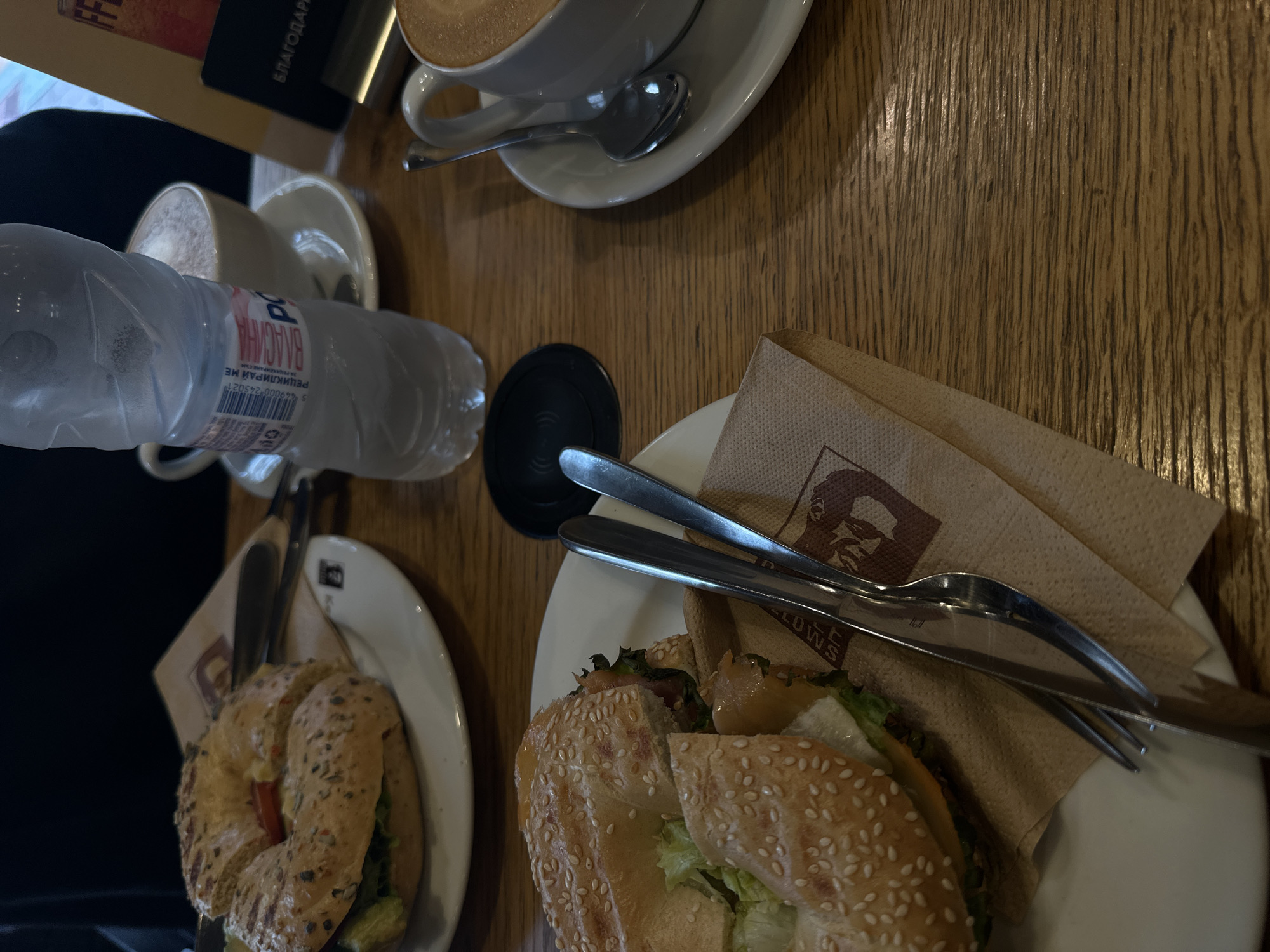 The image depicts a tabletop scene in what appears to be a cafe in Sofia, Bulgaria. The tabletop has a wood grain texture. Visible are two plates, each holding a bagel sandwich. There is a partially visible coffee drink in a white ceramic cup to the left, with a second cup and coffee drink with an associated spoon above the bagel. A water bottle is placed near one of the cups. Napkins are arranged under cutlery, and the restaurant is Caffe & Bellows, as shown on one of the napkins. The scene suggests a meal being consumed or just concluded at this location.