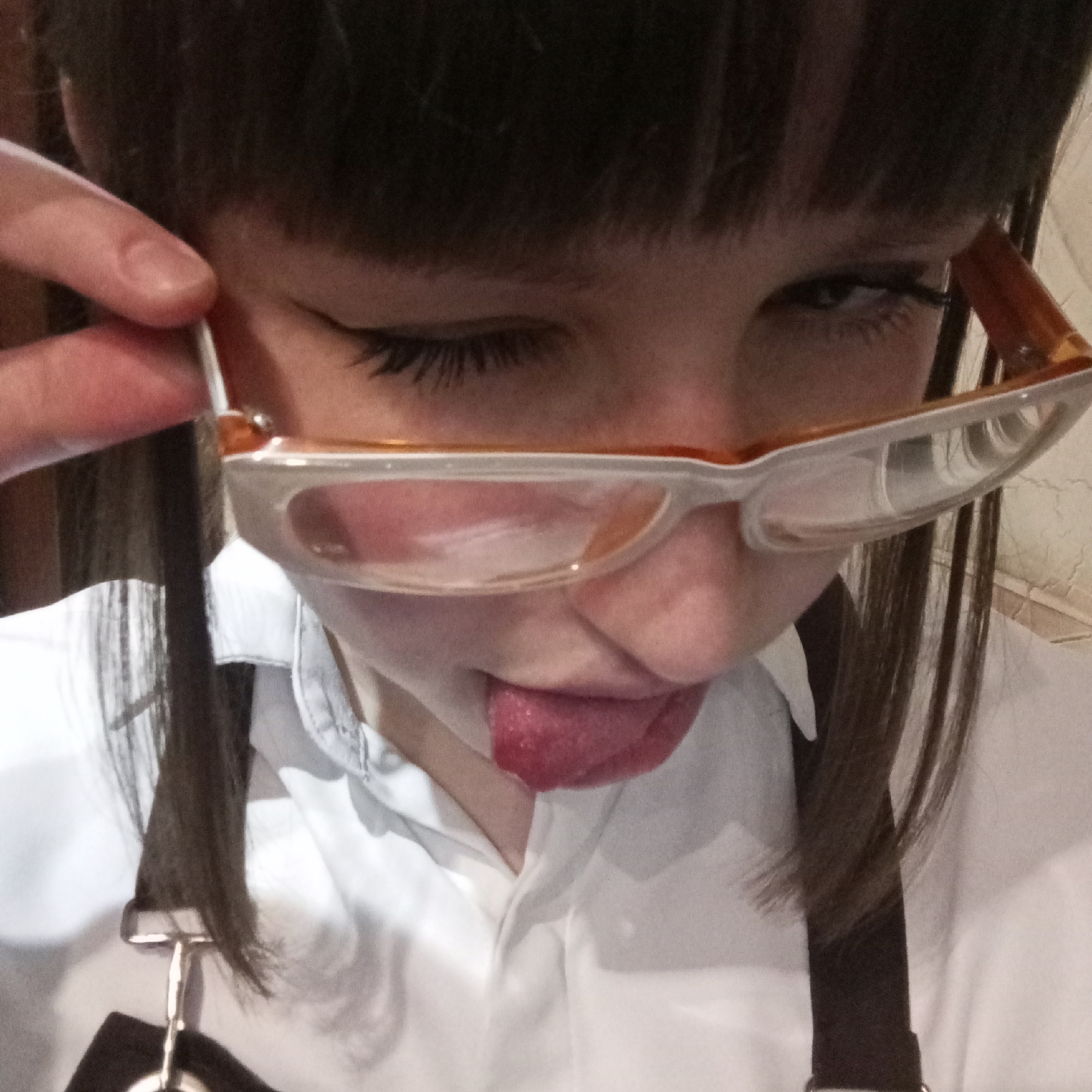 A person is holding their glasses while sticking their tongue out, winking one eye. They have dark hair and are wearing a white collared shirt. There is no discernible information in the image to determine its location in Chaikovsky, Russia.