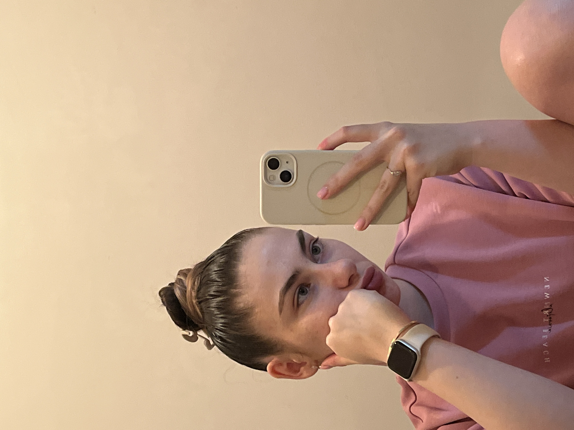 The image depicts a young woman taking a selfie, likely in a mirror, as her smartphone is visible in her hand. The image is oriented with the subject's head to the left and body extending to the right, but is described as if rotated to an upright position.
The woman has dark hair pulled back into a slick bun, secured with a light-colored hair accessory. Her face is in profile, looking slightly upwards and to the right, with a pensive or thoughtful expression. Her left hand (appearing as her right in the rotated image) is resting near her chin and mouth, with her index finger lightly touching her lips.
She is wearing a light pink long-sleeved t-shirt or sweatshirt. On the right side of her chest, reversed text is visible, which reads "NEW HAVEN BEACH". On her right wrist (appearing as her left in the rotated image), she wears an Apple Watch with a light-colored band and a dark screen, along with a thin gold-toned bracelet. A ring with a small, possibly diamond, stone is visible on the ring finger of her left hand, which holds the phone.
She is holding a light-colored smartphone, likely an iPhone, in a matching light beige or cream case. The back of the phone shows a dual-camera setup and a subtle circular MagSafe ring outline. The background is a plain, light beige or cream-colored wall, indicating an indoor setting. The overall atmosphere is calm and introspective.