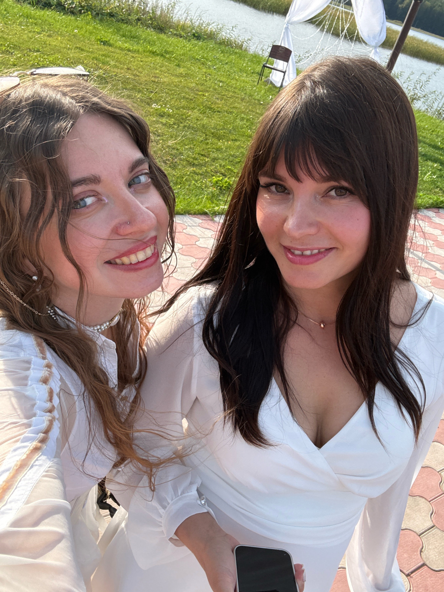 Two women are taking a selfie outdoors. The woman on the left has wavy brown hair and blue eyes. She is wearing a white top with ruffles and a pearl necklace. The woman on the right has straight dark brown hair and brown eyes. She is wearing a white dress. The background shows a grassy area and a body of water. There is a white canopy structure and a chair visible in the distance. The ground in the foreground is paved with reddish-pink and light gray tiles. The image appears to have been taken on a sunny day. The specific location of Пакли, Russia is not identifiable from the visual information provided in the image.