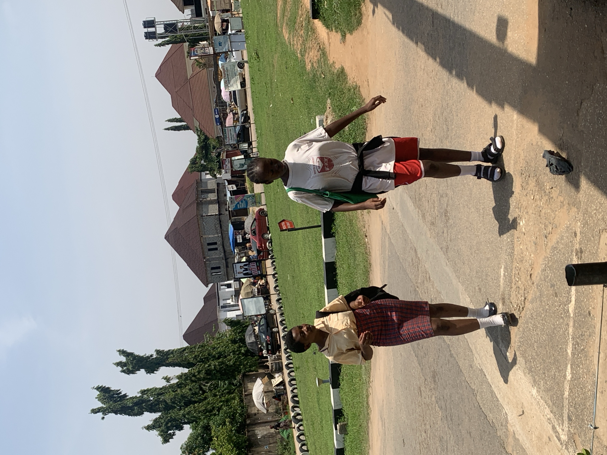 Two young females, appearing to be students, walk on a paved path in a semi-urban environment. The individual on the left wears a tan collared shirt and a red plaid skirt, with white socks and black shoes. A backpack is slung over her left shoulder. The individual on the right is dressed in a white t-shirt with a graphic design, red athletic shorts, white socks, and black sandals. She carries a green tote bag over her left shoulder and has a dark belt tied around her waist.
The background shows a grassy area bordered by a black and white striped curb. Beyond this, a street is visible with several parked cars and buildings under construction or recently built, characterized by brown, angular roofs. In the distance, more complex structures and a water tower can be seen. The overall setting suggests a location within Abuja, Nigeria, possibly near a school or residential area, under a clear, bright sky. The ground is asphalt and dirt, with a noticeable shadow cast by the individual on the right.