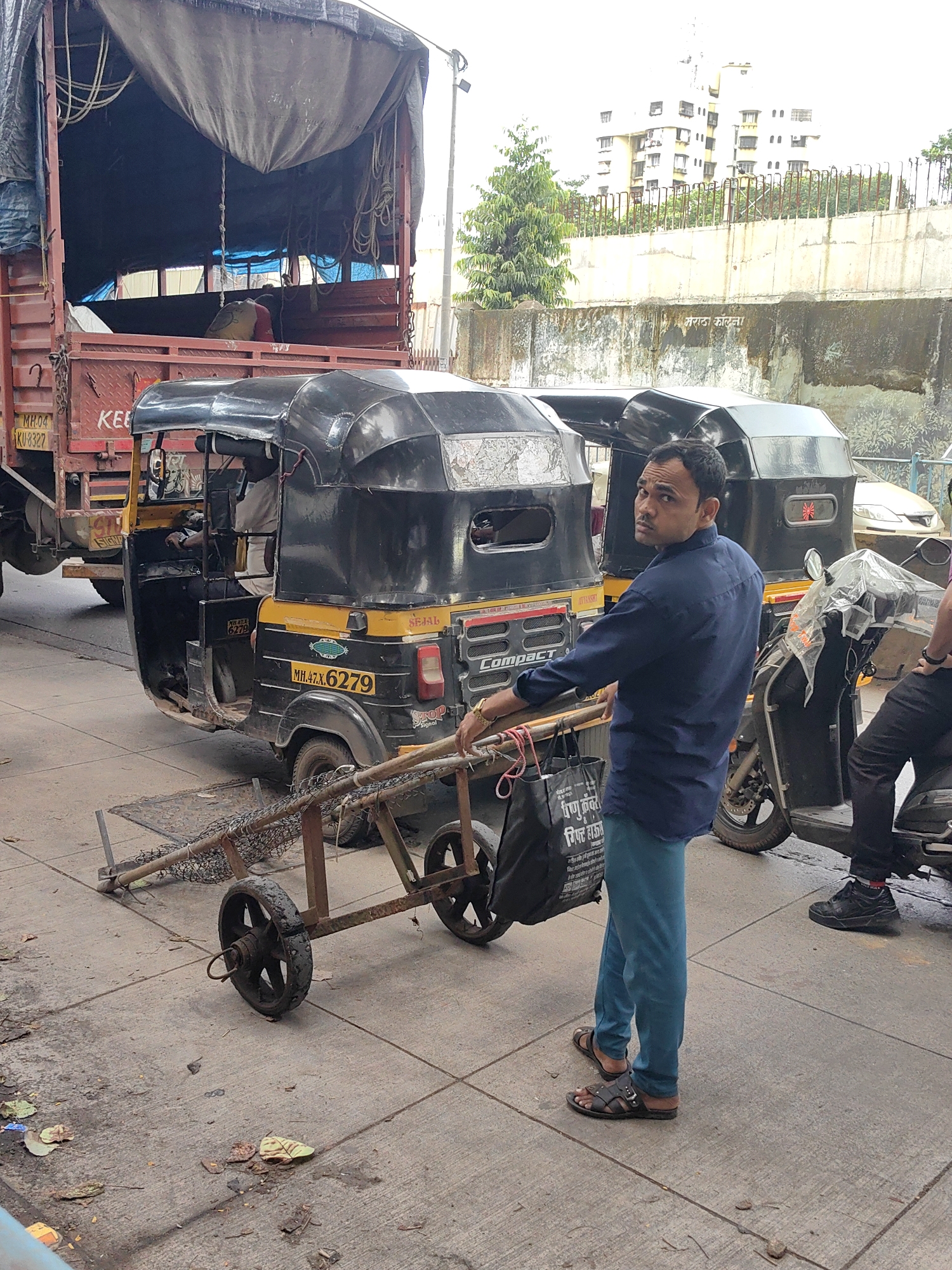 The image depicts an outdoor scene in Mumbai, India. In the foreground, a man wearing a blue shirt and turquoise pants is standing next to a two-wheeled cart with a mesh basket. He is looking towards the camera. The cart appears to be a utility cart, possibly for carrying goods. Behind the man, a black and yellow auto-rickshaw is parked, identified by the license plate MH.47.X.6279. Another auto-rickshaw is partially visible to the right. In the background, a large truck with a tarp-covered cargo is parked, and beyond that, a multi-story building and a concrete wall are visible. The ground is paved with concrete tiles, and there are some scattered leaves and debris. The overall impression is of a street scene in an urban environment, likely a busy area with transportation and commercial activity.