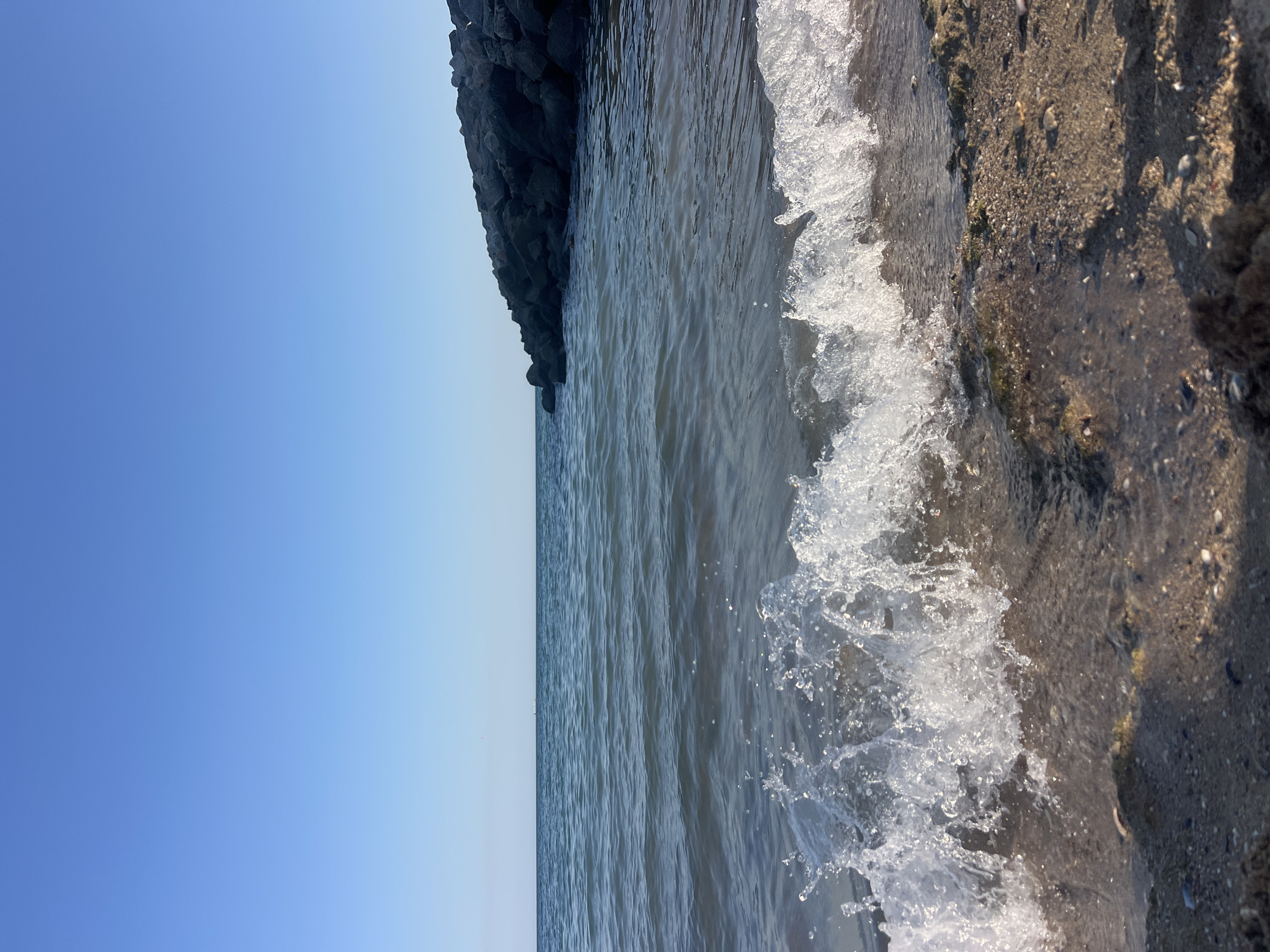 A low-angle shot captures a wave crashing on a sandy beach under a clear blue sky. The water is a deep blue, with ripples and foam from the breaking wave in the foreground. To the right, a breakwater made of large, dark rocks juts out into the sea. The sand on the beach is a light brown, speckled with small shells and pebbles, and appears wet from the receding wave. Shadows of objects, not visible in the frame, are cast across the sand. The horizon line is clearly defined where the water meets the sky. The image is taken at eye-level with the wave as it breaks on the shore.