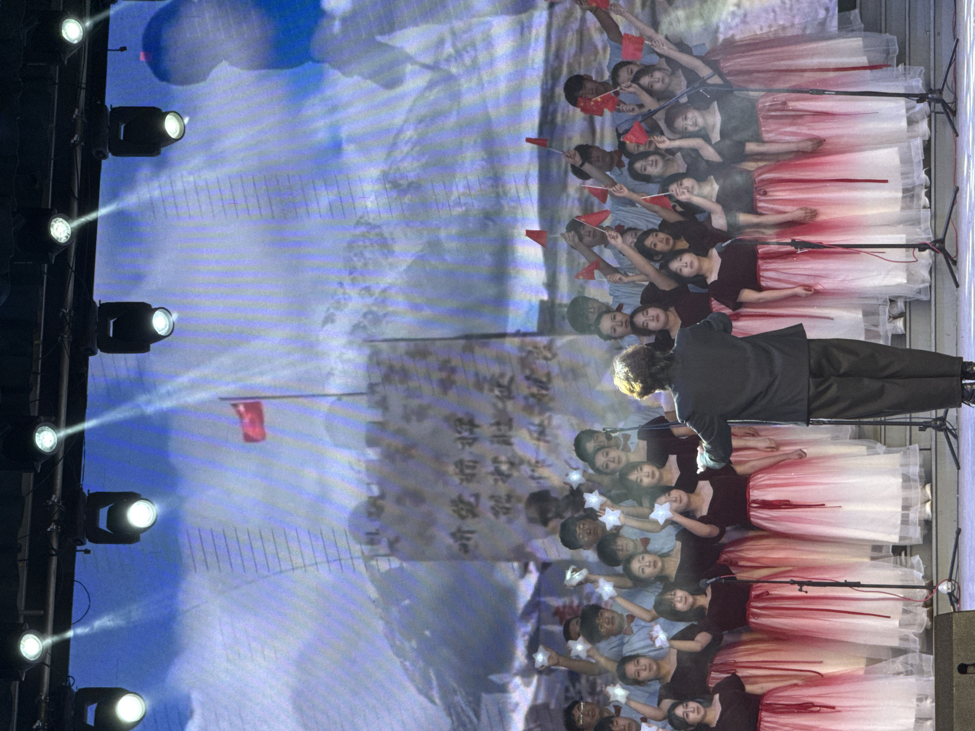 This image captures a stage performance, likely a choral or patriotic event, taking place indoors or at night under artificial lighting. The scene is dominated by a large group of performers arranged in rows on a stage.
At the forefront, with his back to the camera, stands a man in a dark suit, possibly a conductor or lead performer. Behind him, several rows of young women are visible, wearing dark tops and distinctive skirts that gradient from dark red at the top to white at the bottom, creating a flowing effect. Many of these women have microphones positioned in front of them on stands.
Behind the female performers, a group of young men or boys in light blue shirts can be seen. Many of the performers, both men and women, are holding small red flags adorned with yellow stars, consistent with the national flag of China. Some of the male performers are also holding white, star-shaped objects that appear to be illuminated.
The background features a large projection screen displaying an image that resembles mountainous or cloud-filled terrain, with faint Chinese characters visible on its surface. Bright spotlights are positioned along the left edge of the frame, casting strong beams of light onto the stage and performers. Stage equipment or speakers are partially visible on the right.
The overall atmosphere suggests a formal and perhaps celebratory or nationalistic performance. The presence of Chinese flags and characters on the screen, along with the given location context of Guta, China, reinforces this interpretation.