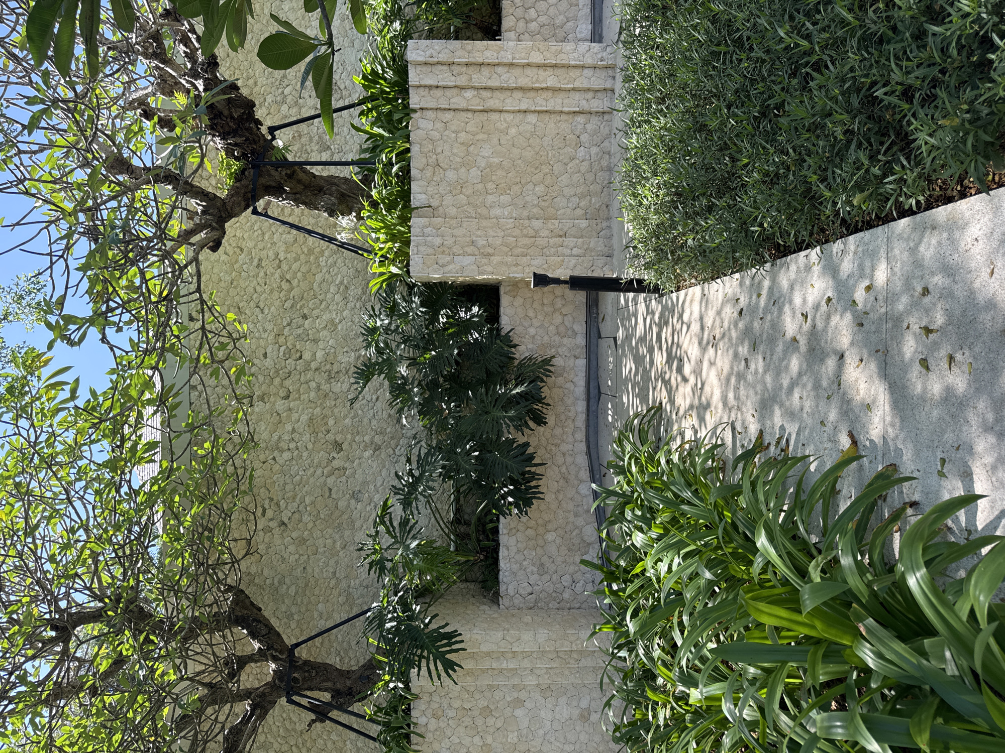 The image depicts a stone-paved pathway bordered by various plants and trees, set against a stone wall. The path leads upward via steps constructed from the same stone material as the wall. A tree with sparse foliage is situated on the left, supported by a metal framework. On the right, dense green shrubbery lines the path, casting shadows on the paved surface. Additional foliage peeks out from crevices within the stone wall. The location is Kuta Selatan, Indonesia.