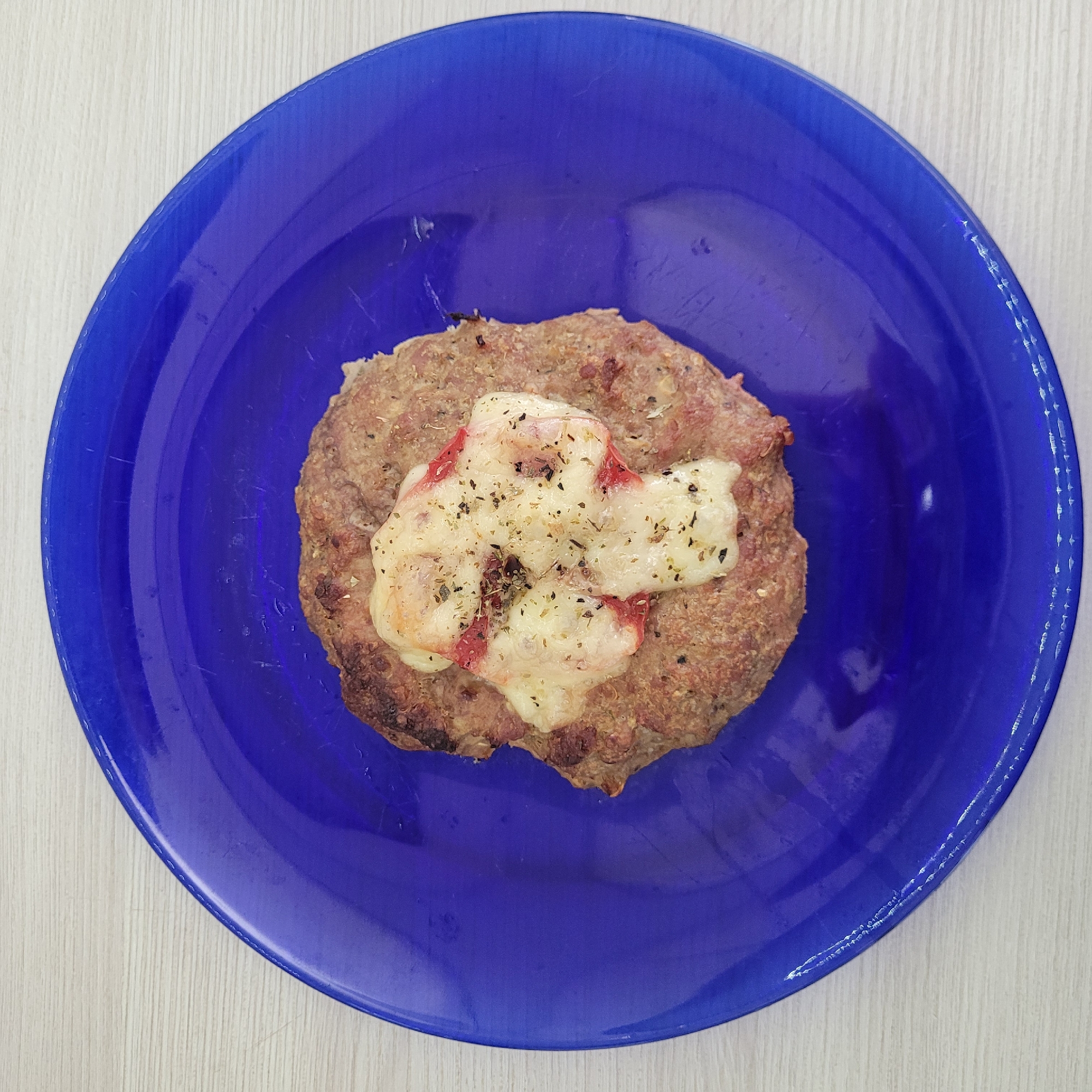 A close-up, overhead shot captures a single, cooked hamburger patty topped with melted cheese and red ingredients, possibly tomato or pepper, with dried herbs sprinkled on top. The patty rests on a vibrant blue, round plate. The plate itself is placed on a light-colored, textured surface that appears to be a wooden table. The lighting is bright and direct, casting subtle shadows. The image does not provide any information that would allow for the determination of its location in Jūrmala, Latvia, or any other specific city.