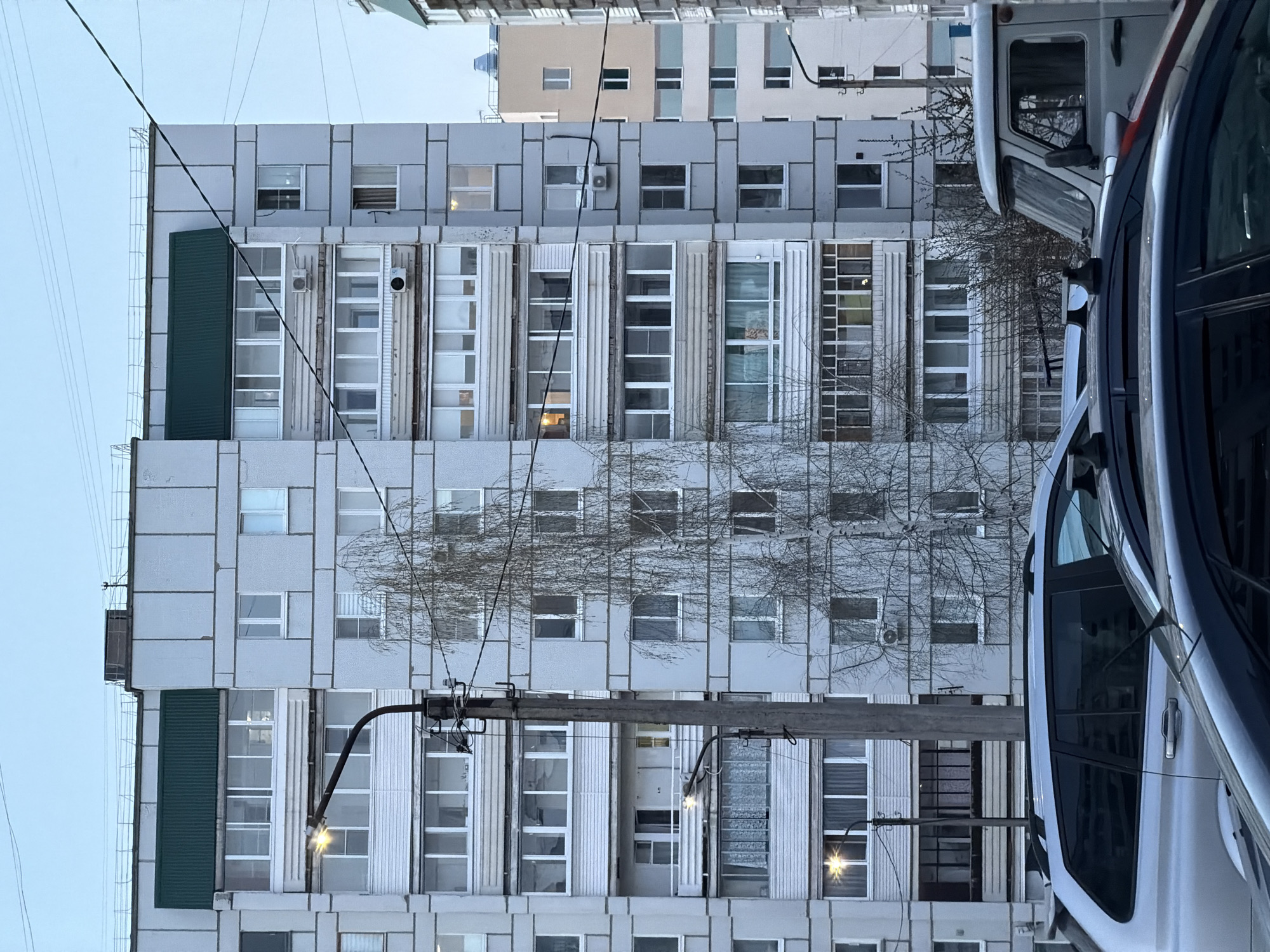 A building with rows of windows is featured in this photo, with a car partially visible on the right side of the frame. The building has a gray facade, with visible architectural details. The windows are a mix of clear and frosted glass. A leafless tree stands in front of the building. A street light is positioned in the lower part of the image. The photo's location is the city of Chaikovsky, Russia.