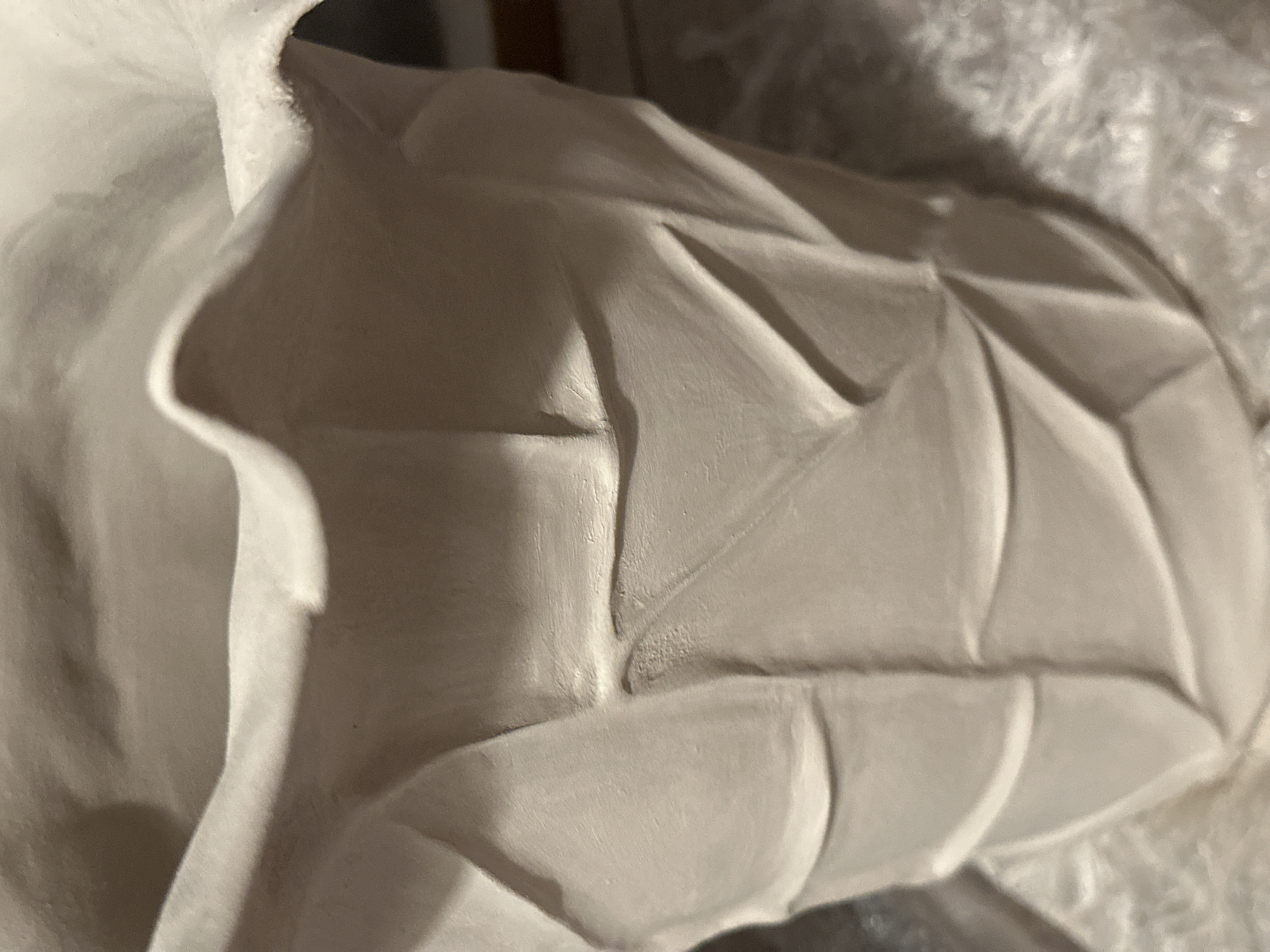 A close-up, low-angle, eye-level shot shows the textured surface of a light-colored, perhaps unglazed, ceramic vase. The vase has a wide, ruffled opening at the top that curves inward. The body of the vase is decorated with a geometric pattern of folded or creased sections, creating a faceted appearance. The lighting casts shadows that emphasize the three-dimensional texture of the ceramic. The background is out of focus and appears to be a light-colored, possibly textured surface, with some darker areas suggesting a surrounding environment. The location in Nevinnomyssk, Russia, is not discernible from the image. There are no people or animals present.