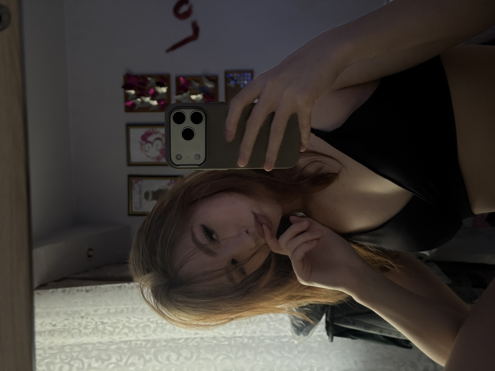 A young woman with red hair is taking a selfie in a mirror. She is holding a cell phone with a gray case, and her right hand is near her mouth. She is wearing a black top. The background includes a bed with a patterned bedspread, framed pictures on the wall, and a piece of red lettering above the frames. The location in the city of Noviy, Russia is indeterminable.