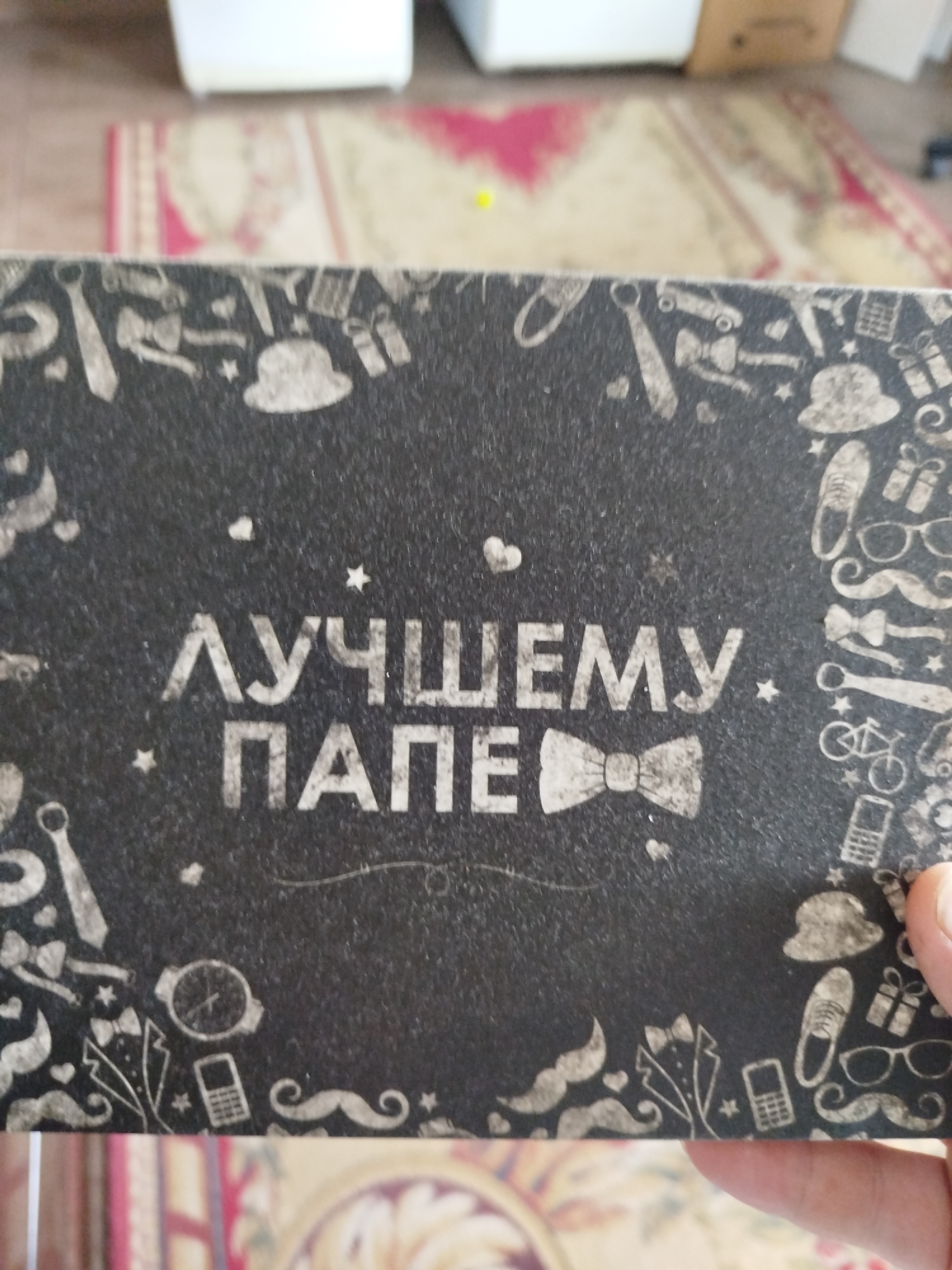 The image shows a close-up of a black gift box with white decorative patterns and text. The text, in large white Cyrillic letters, reads "ЛУЧШЕМУ ПАПЕ", which translates to "To the best dad". The decorative elements include images of bow ties, hats, ties, shoes, glasses, mustaches, a bicycle, a gift box, a watch, and a mobile phone. The box is resting on a surface that appears to be a rug with a patterned design. The background is blurred, but suggests an indoor environment with hints of furniture. The image is taken from a slightly angled perspective, and a person's hand is visible on the right side of the frame, holding the box. The lighting suggests it is daytime, and the overall scene appears to be domestic. The specific location of Крывасельскі сельскі Савет, Belarus, is not visually ascertainable from the image itself.