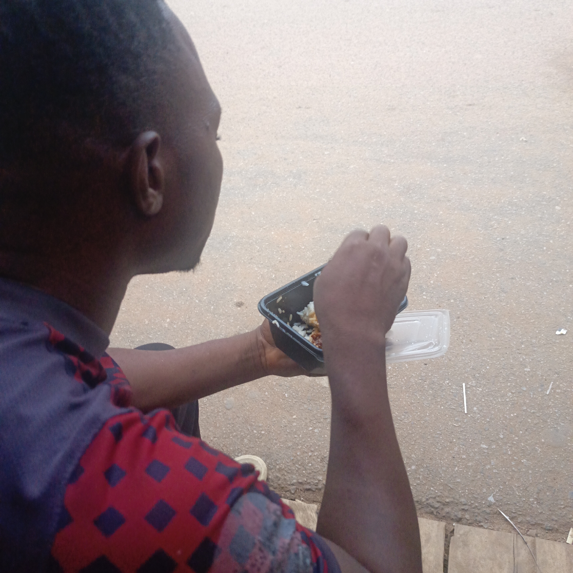A person is eating from a black plastic container with a clear plastic lid in an outdoor setting, likely in Akwanga, Nigeria. The person is facing away from the camera, and the location appears to be a street or sidewalk with scattered pieces of trash. The person's clothing has a pattern of red with blue squares. The food in the container is rice and possibly another item, and the person is using a utensil. The ground is a light brown or tan color and appears to be asphalt or concrete.