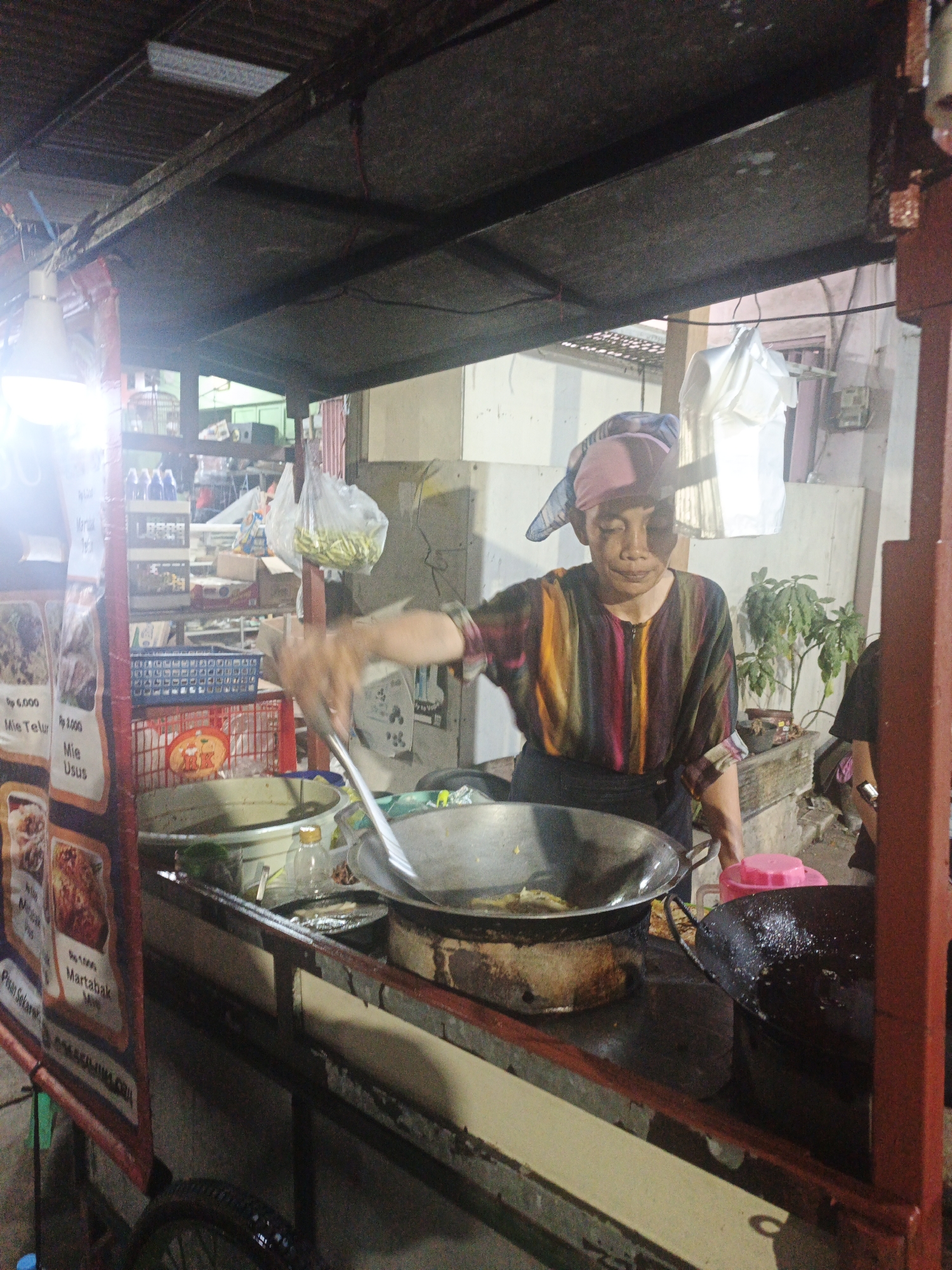 The image depicts a street food stall, likely in the city of Surabaya, Indonesia, with a vendor actively cooking. The stall is covered by a corrugated metal roof, supported by red metal framing. A bright fluorescent light is visible on the ceiling of the stall. The vendor, wearing a headscarf and a colorful striped shirt, is using a large metal spatula to stir food in a frying pan. To the right of the frying pan, another dark-colored pot or pan is present, also containing food. Behind the vendor, shelves are stocked with various items, including what appear to be plastic containers and bags of food. A sign with Indonesian text and images of food is affixed to the left side of the stall. A bicycle wheel is visible at the bottom left of the image, suggesting the stall is mobile or parked. The overall environment appears to be an outdoor market or street setting, with a slightly cluttered and utilitarian aesthetic typical of street food vendors.