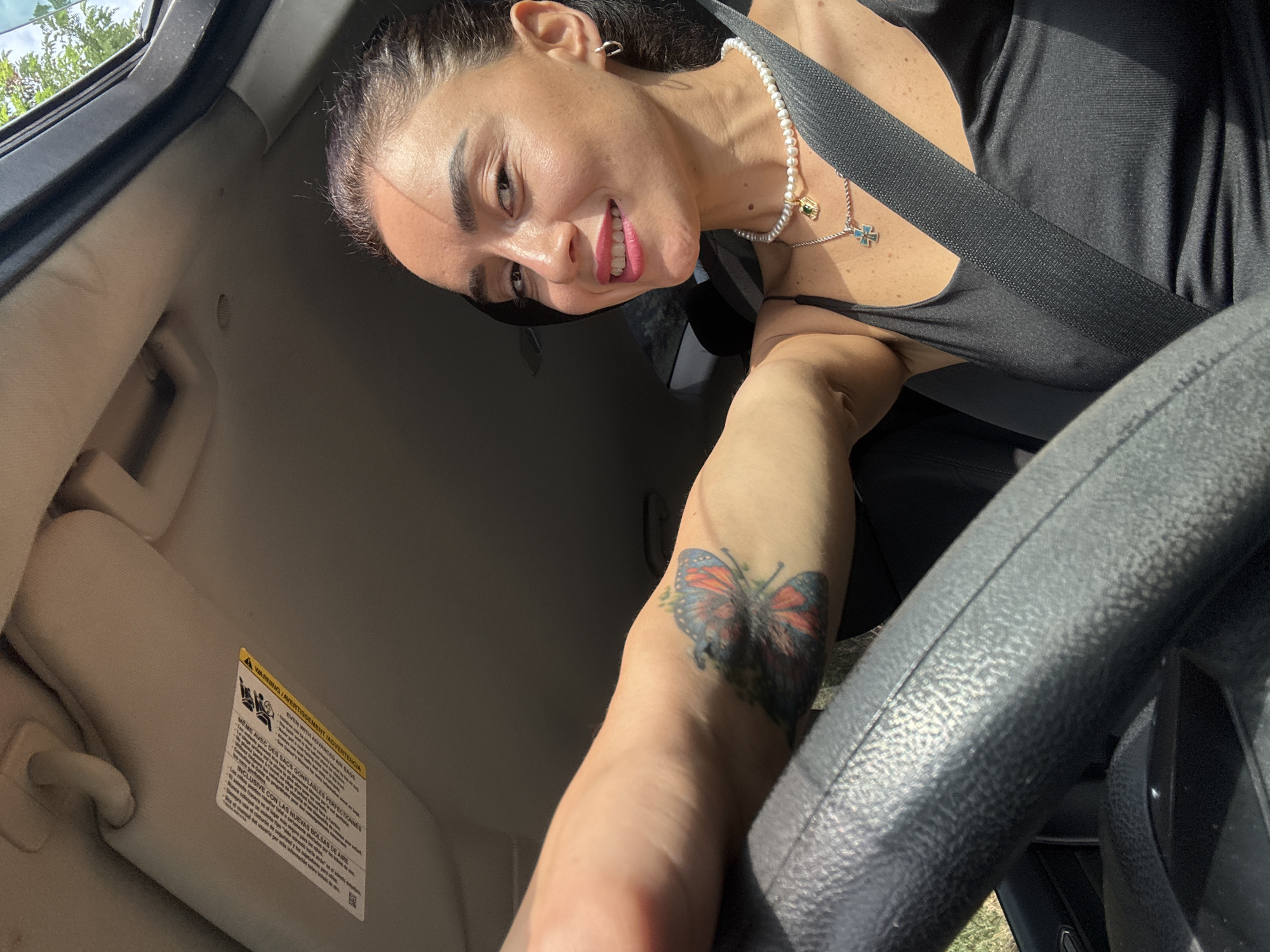 A woman is pictured in the driver's seat of a car, with her left arm extended as she holds the steering wheel. She is wearing a black tank top and a pearl necklace with two pendants, one of which is a green gemstone and the other a cross. A seatbelt is fastened across her chest. A tattoo of a butterfly is visible on her upper left arm. The car appears to be in motion, as indicated by the blurred background of trees and foliage, suggesting an outdoor setting, possibly on a road outside of Tbilisi, Georgia. The interior of the car shows the sun visor and part of the dashboard.