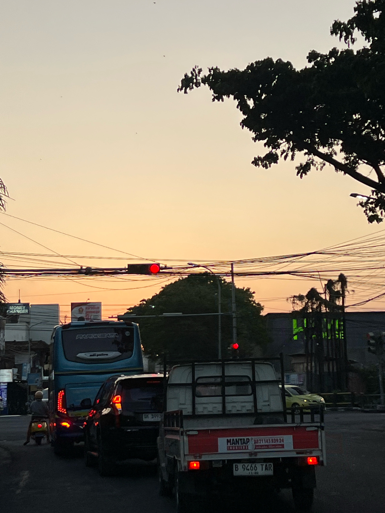 The image captures a street scene in Kuta, Indonesia, during sunset. A traffic light shows a red signal, indicating a halt. In the foreground, a white pickup truck is stopped, displaying a license plate that reads "B 9466 TAR". To its left, a blue bus with "PARIWISATA" written on its back is also stationary. A person on a yellow scooter is visible to the left of the bus. The background features buildings, trees, and a sky with a warm orange hue. Overhead, a dense network of electrical wires is prominent. The overall atmosphere suggests a moment of traffic congestion in a bustling urban environment.