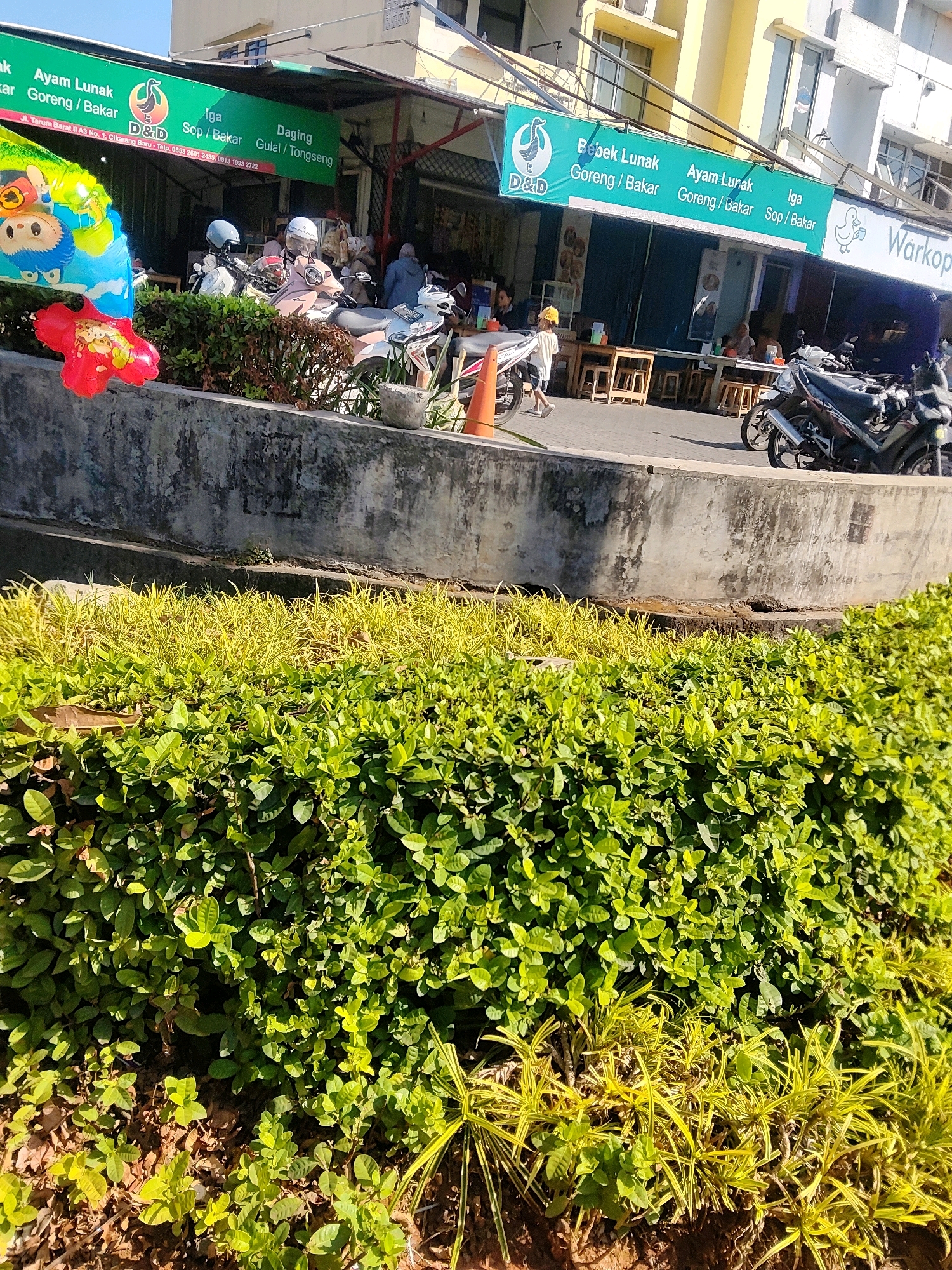 The image shows a street scene in Cibatu, Indonesia, likely outside a food establishment. A green awning with the text "Ayam Lunak Goreng / Bakar" and "D&D" is visible, indicating a restaurant specializing in chicken dishes. Next to it, another awning displays "Bebek Lunak Goreng / Bakar," "Ayam Lunak Goreng / Bakar," and "Iga Sop / Bakar," suggesting a menu offering duck, chicken, and ribs. The word "Warkop" is also visible, a common term for small Indonesian eateries. Several motorcycles are parked along the side of the road. In the foreground, lush green bushes and yellow ornamental grasses are planted. A concrete barrier separates the road from the vegetation. A few people are seated at tables under the awnings, and one person is walking away from the camera. Children's balloons are present on the left side of the image, suggesting a festive atmosphere or a nearby event. The buildings in the background appear to be multi-story structures. The sky is clear and blue, indicating a sunny day.