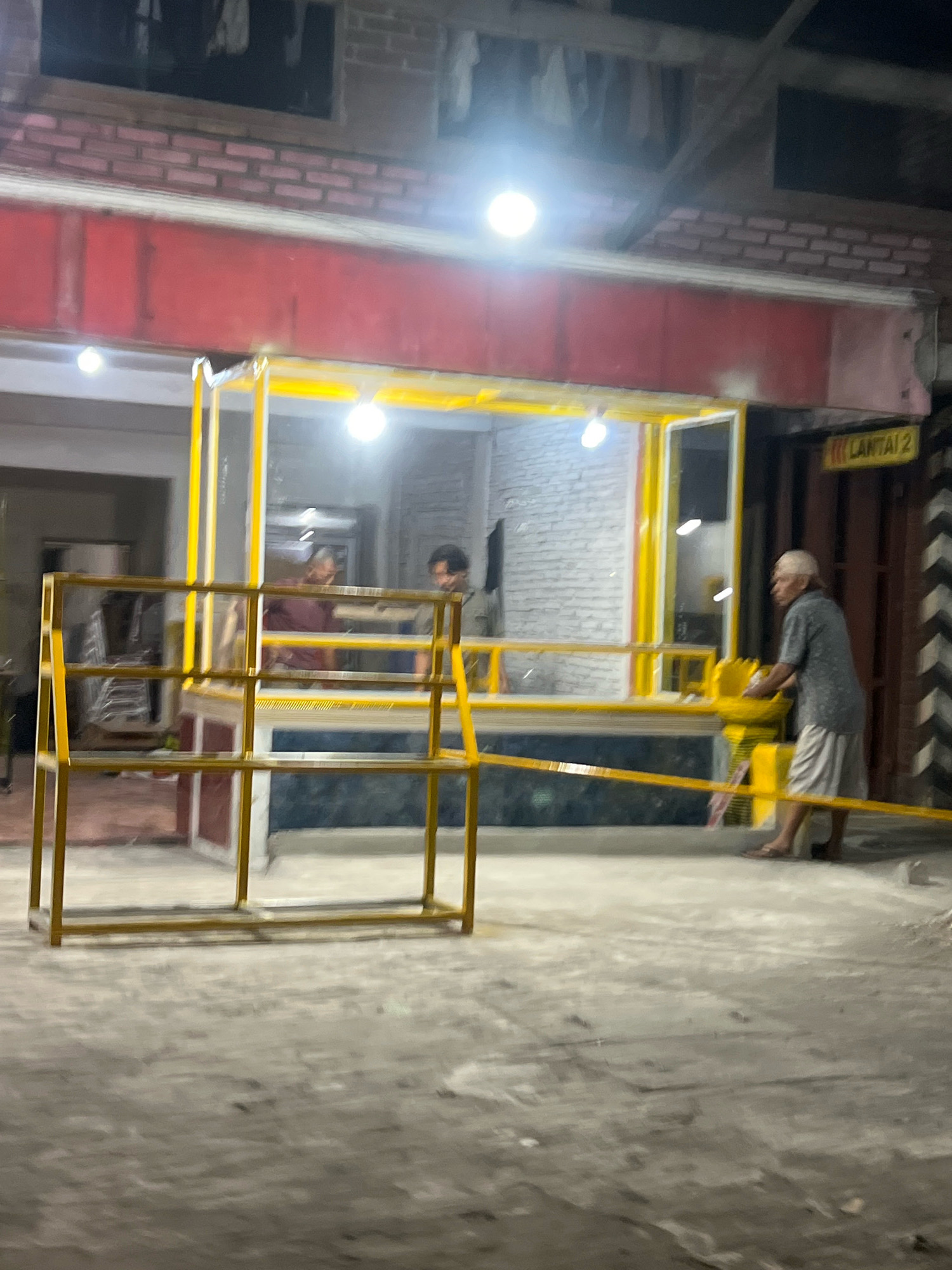 The image depicts an outdoor scene at night, likely in a public space. A small, open-fronted kiosk with a yellow frame and red exterior is visible, illuminated by bright lights. Inside the kiosk, two men are present; one appears to be serving or interacting with the other. To the right of the kiosk, an older man wearing a short-sleeved shirt and light-colored pants is standing and looking towards the kiosk. He is partially obscuring a yellow sign on a wooden door which reads "LANTAI 2". The ground appears to be a concrete or paved surface. A yellow metal structure, possibly a barrier or display rack, is in the foreground. The overall environment suggests a commercial or service area. The location is identified as Secang, Indonesia.