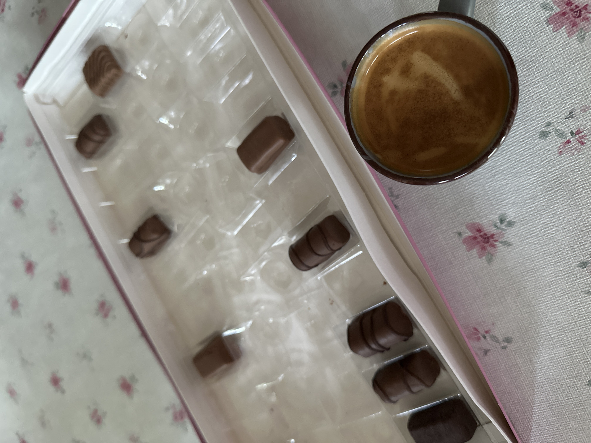 The image shows a box of chocolates and a cup of coffee. The box is pink and white, and it contains several different types of chocolates. Some of the chocolates are missing from the box. The cup of coffee is dark brown and has a layer of crema on top. The background is a tablecloth with a floral pattern. This image was taken in Garmisch-Partenkirchen, Germany.