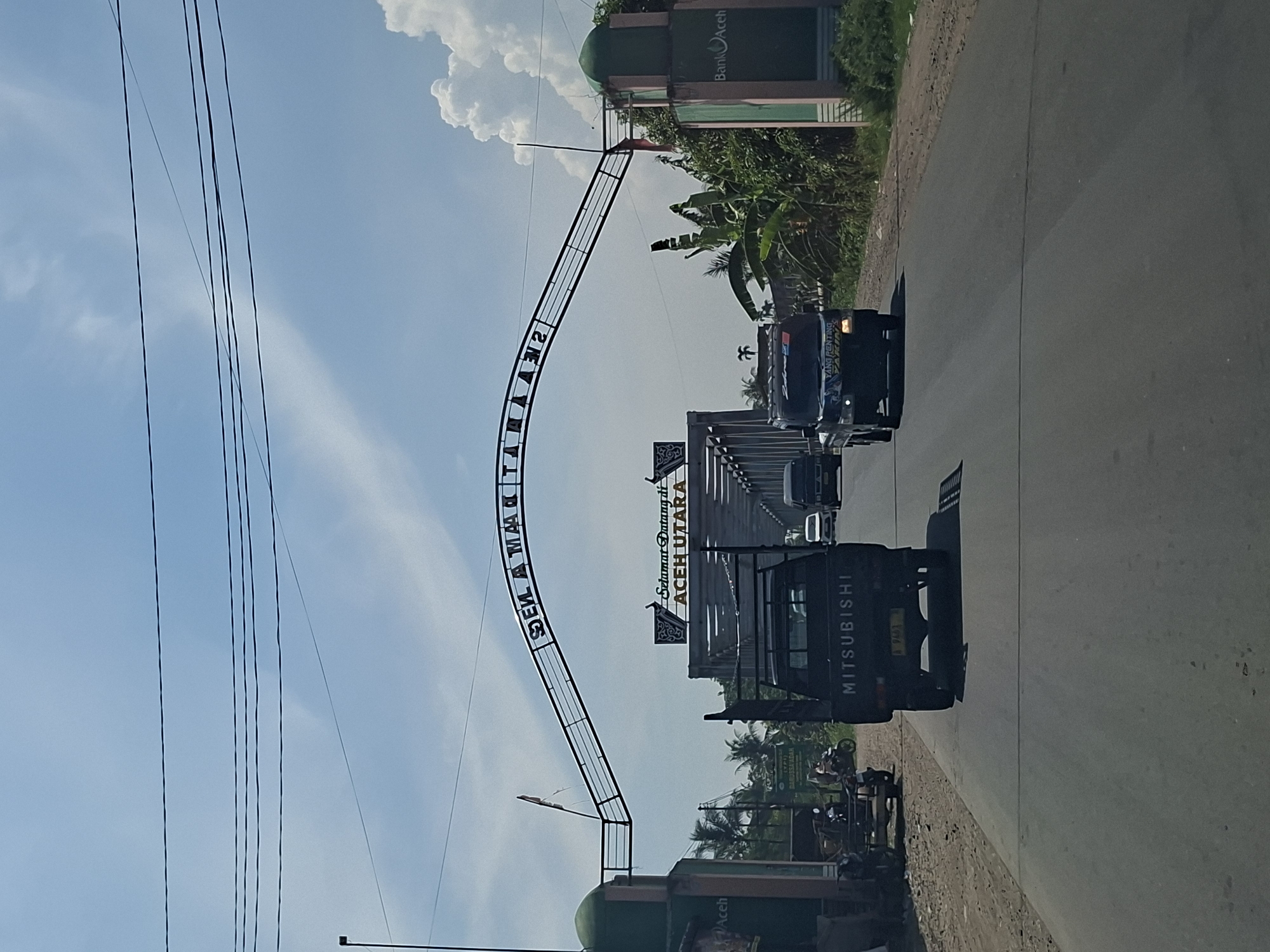 This is a photograph taken from a moving vehicle in Teupinsiron, Indonesia, looking towards a bridge and an archway spanning the road. The archway displays the words "SELAMAT DATANG DI ACEH UTARA" in black lettering against a white background, indicating a welcome to North Aceh. Two trucks are visible on the road; the front truck is a black Mitsubishi pickup truck with a license plate that reads "B 9453 PA". Behind it, another black pickup truck is driving away from the camera. The environment features a clear blue sky with scattered clouds, power lines overhead, and vegetation including palm trees and banana plants. A building with "Bank Aceh" signage is present on the right side of the road, and another building with green and pink accents is on the left. Several motorcycles and a cart are parked on the side of the road near the left building.