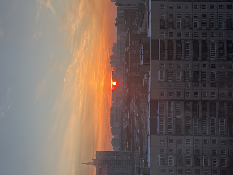 A vertical view showcases a city skyline during sunset. The sky transitions from light blue to fiery orange, with streaky clouds adding texture. The sun appears as a bright red orb, partially obscured by the buildings. Tall apartment buildings with repeating window patterns dominate the right side, contrasting with the fading sunlight. Other urban structures are silhouetted against the colorful sky.