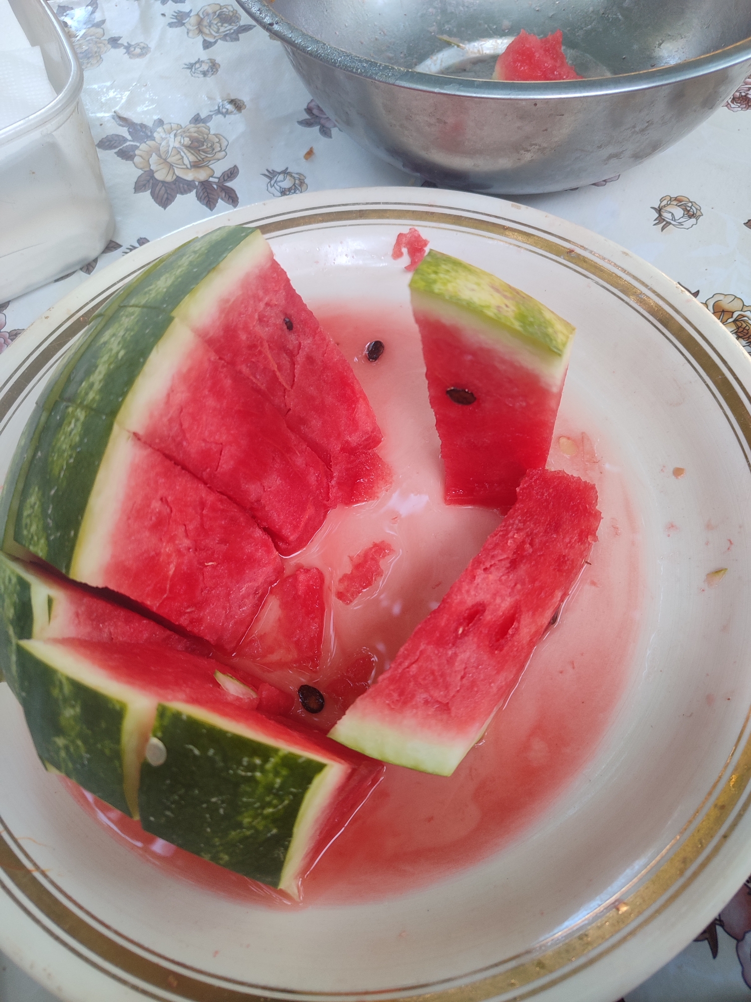 A plate of sliced watermelon is shown in the foreground. The watermelon is red and has black seeds. The plate is white with a gold rim. In the background, a metal bowl with some watermelon scraps is visible. The scene appears to be outdoors on a table with a floral tablecloth.