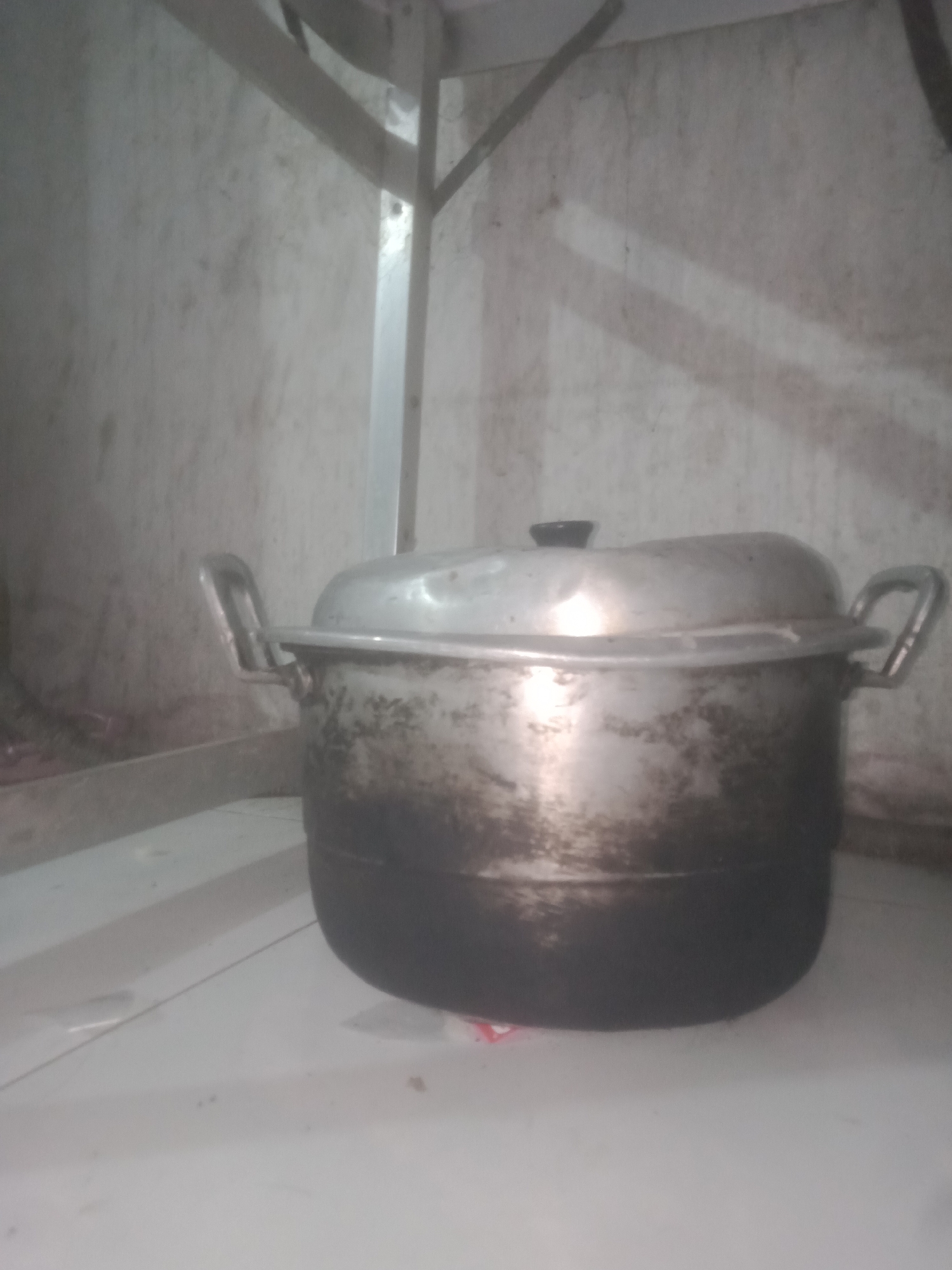 A weathered, aluminum pot with two handles sits on a white surface. The pot is stained with dark soot around its base. It has a domed lid with a black knob. The background appears to be a wall with a metal shelf above it, suggesting it might be in a kitchen or cooking area. The scene is dimly lit, with some shadows and glare on the pot. The location is Badas, Indonesia.