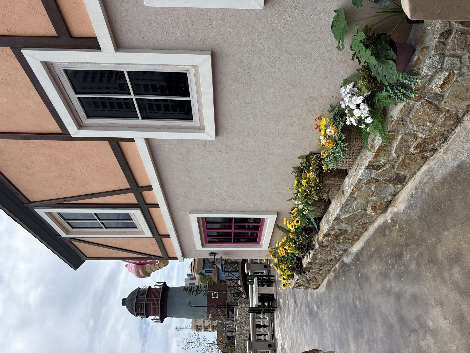 The image shows a street scene in Pattaya City, Thailand. In the foreground, a paved walkway leads past a low stone wall adorned with potted plants and baskets of flowers. The wall is made of rough-hewn stones in varying shades of brown and gray. Behind the wall, a stucco building with a salmon-colored upper facade and dark wood trim features two windows. The window on the right is large and square with dark panes, while the window to its left is smaller and rectangular with a vibrant pink frame. Further in the background, a tall lighthouse with a dark roof and a red railing is visible. Several other buildings, including a blue-colored one and a white multi-story structure, are also present. The sky is overcast.